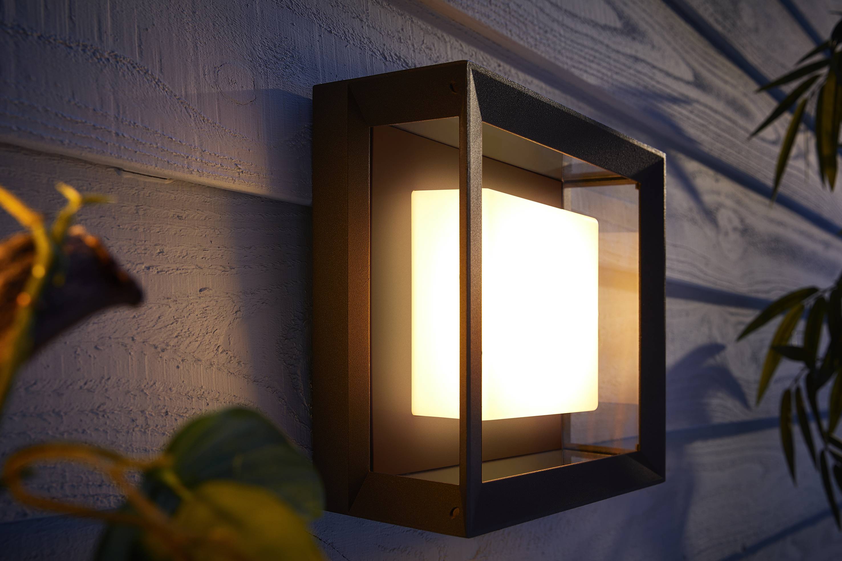 Una moderna applique da parete illumina una parete rivestita di listelli di legno. Foglie di piante sono visibili in primo piano, creando un'atmosfera accogliente.