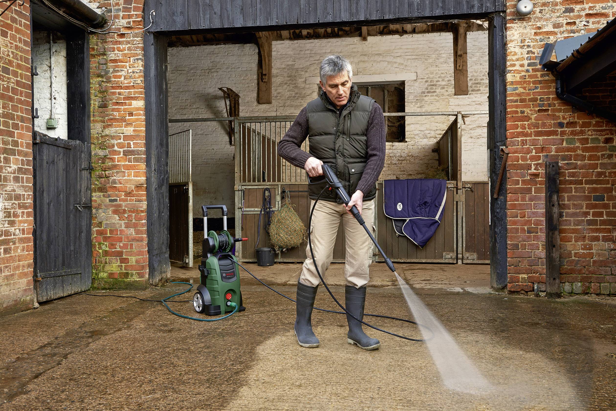 Un uomo sta pulendo un cortile interno con un idropulitrice. Sullo sfondo sono visibili una stalla e attrezzature agricole.