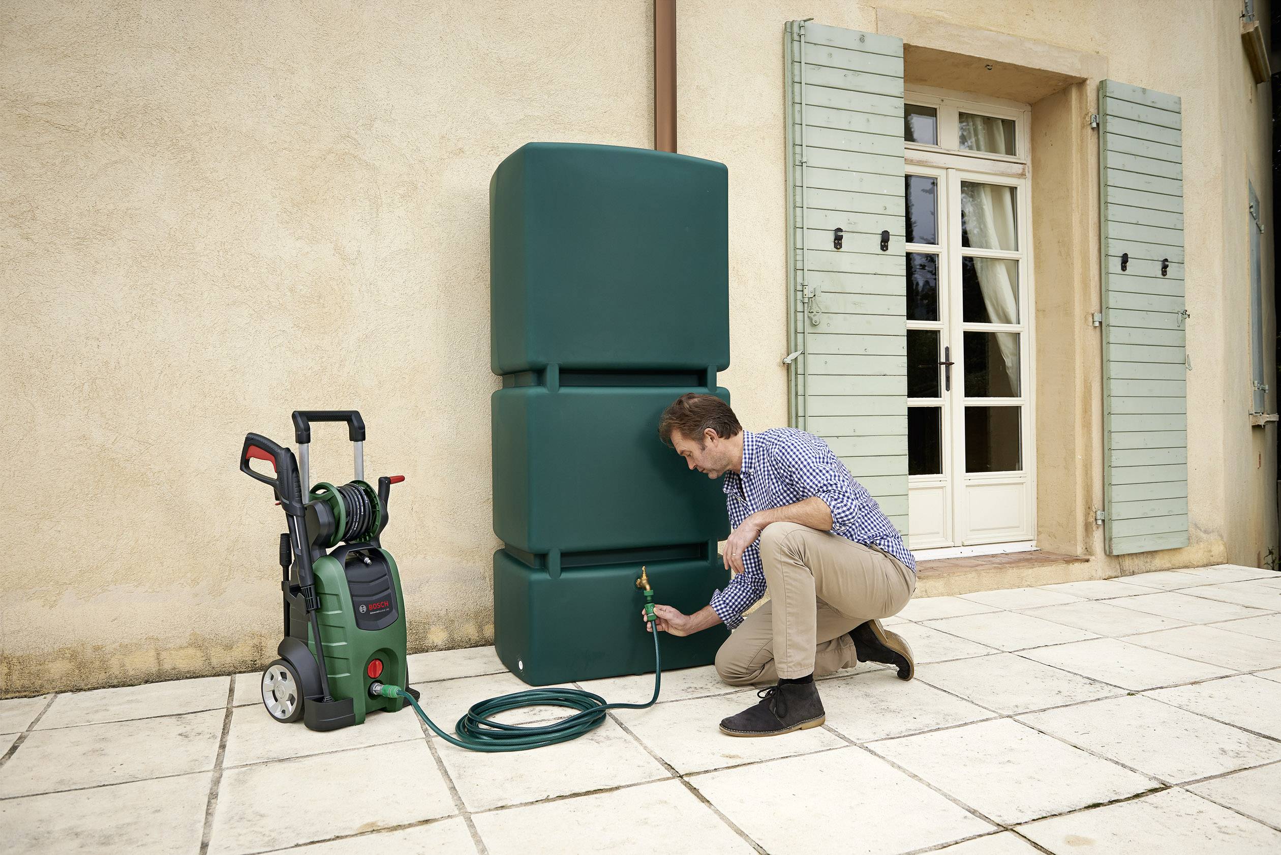 Un uomo è inginocchiato accanto a un serbatoio verde dell'acqua all'aperto e sta fissando un tubo da giardino. Accanto a lui c'è un idropulitrice mobile.