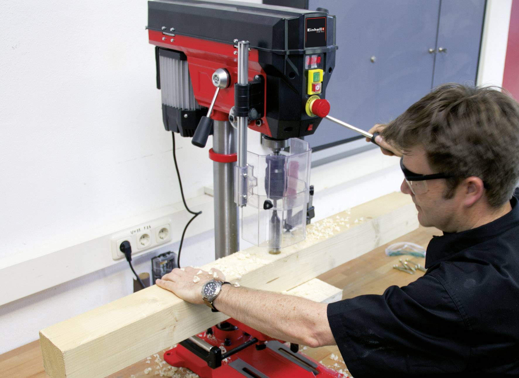 Un uomo utilizza un trapano a colonna per lavorare un grande pezzo di legno in un laboratorio. Sono visibili le precauzioni di sicurezza.
