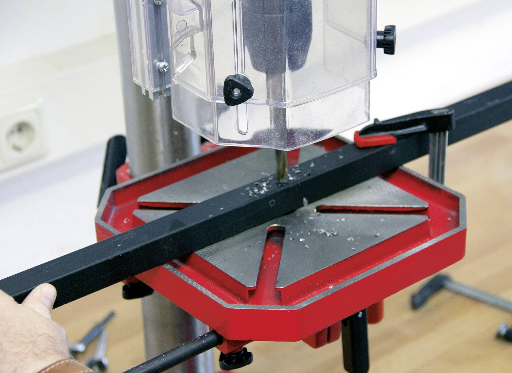 Un pezzo di metallo viene fissato su un supporto per trapano rosso e nero. Un involucro protettivo circonda il trapano, che si trova sopra il pezzo.