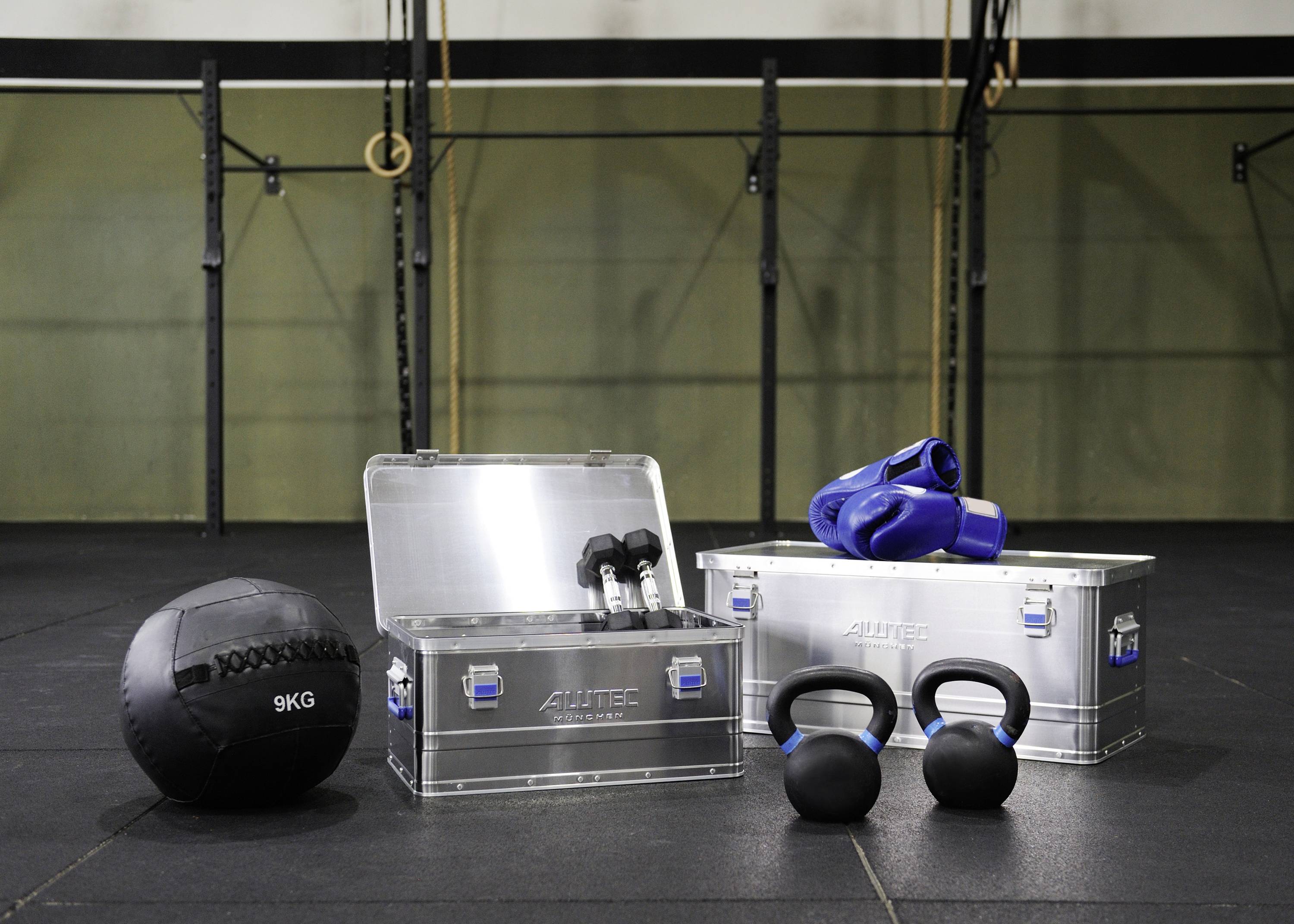 Palestra con attrezzatura CrossFit: palla medica, kettlebell, scatole in alluminio e guanti sportivi sul pavimento davanti a anelli e sbarre per trazioni.