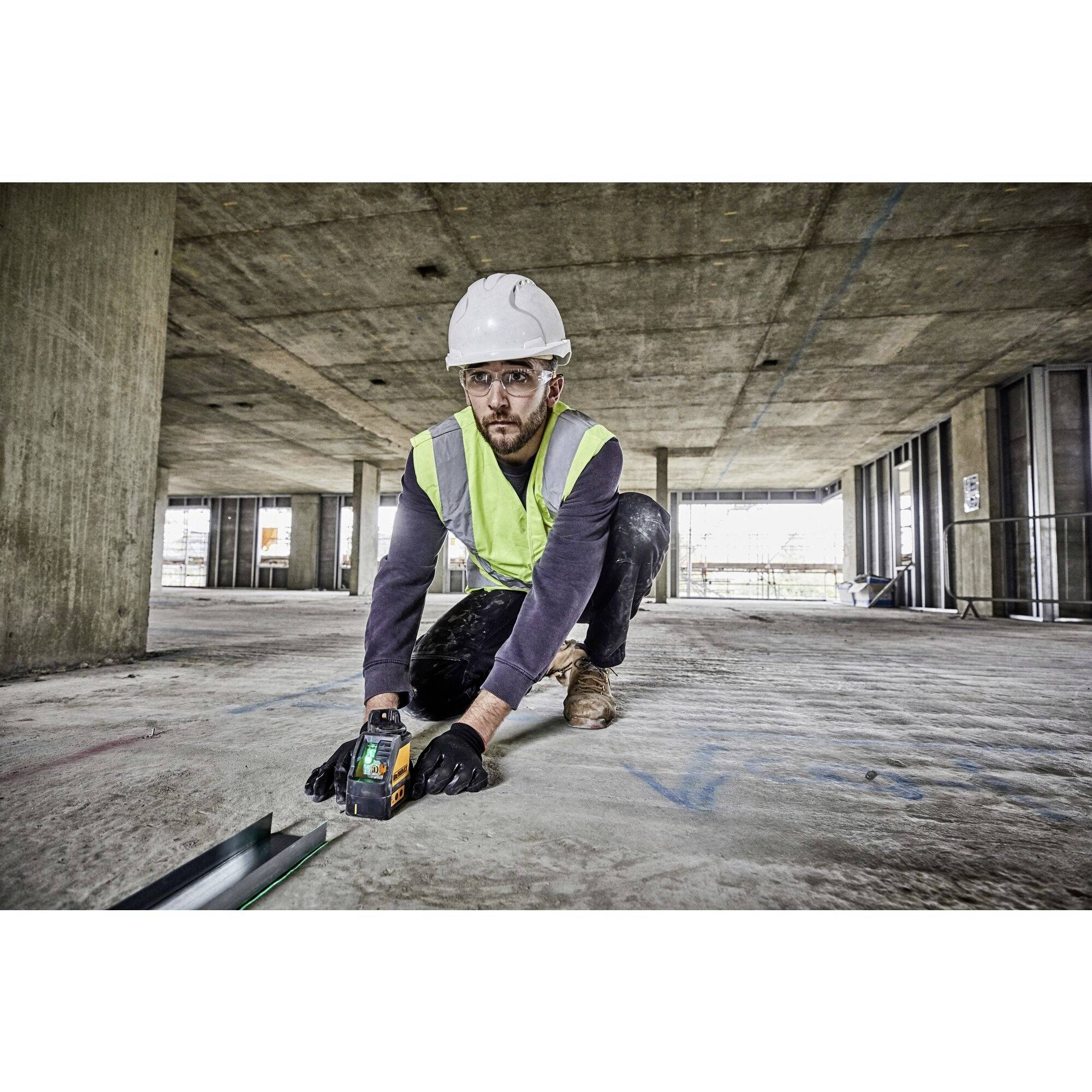 Un operaio edile con giubbotto di sicurezza e casco sta utilizzando uno strumento di misurazione in un cantiere per tracciare segni sul terreno.