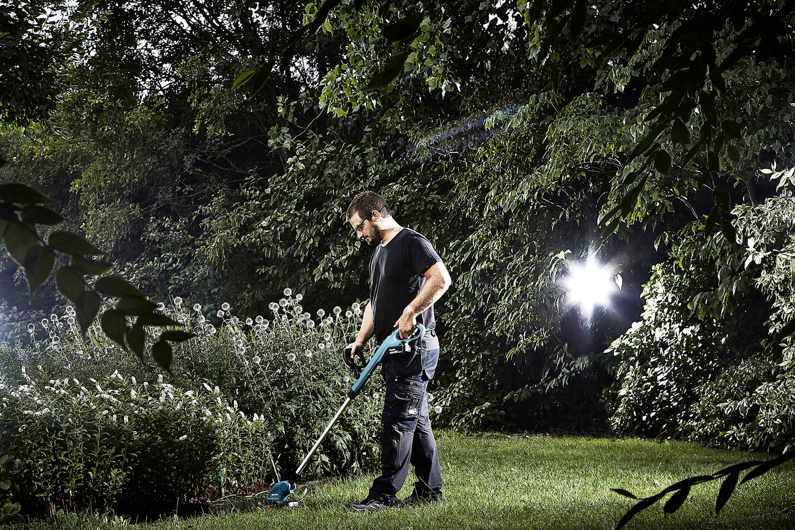 Una persona sta utilizzando un decespugliatore in giardino, circondato da alberi e piante. È una giornata soleggiata, con ombre che si proiettano sul prato.