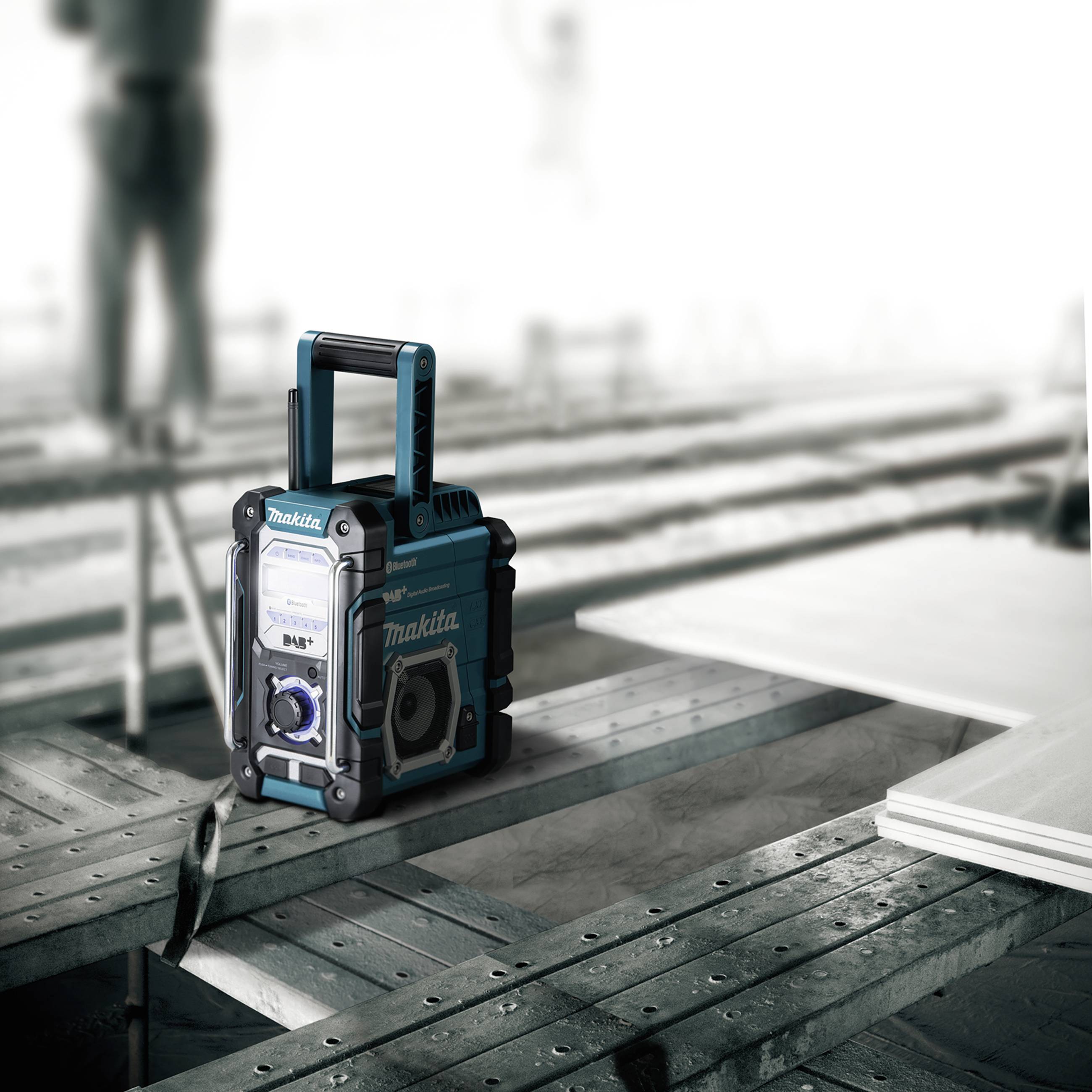 Un radio portatile da cantiere è poggiato su un'impalcatura metallica. Sullo sfondo, figure sfocate lavorano in un cantiere edile.