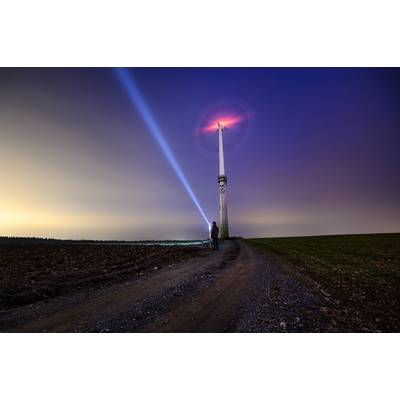 Una persona è in piedi su un sentiero di campagna di notte e illumina con una torcia un mulino a vento illuminato sotto un cielo variopinto.