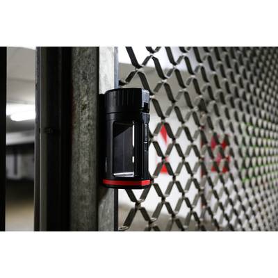 Una torcia rossa e magnetica è attaccata a una griglia metallica in un parcheggio sotterraneo, illuminando leggermente lo sfondo.