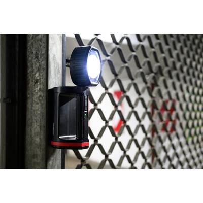 Una telecamera di sorveglianza con luce intensa è fissata a un cancello metallico. Sullo sfondo, un cartello rosso è parzialmente visibile.