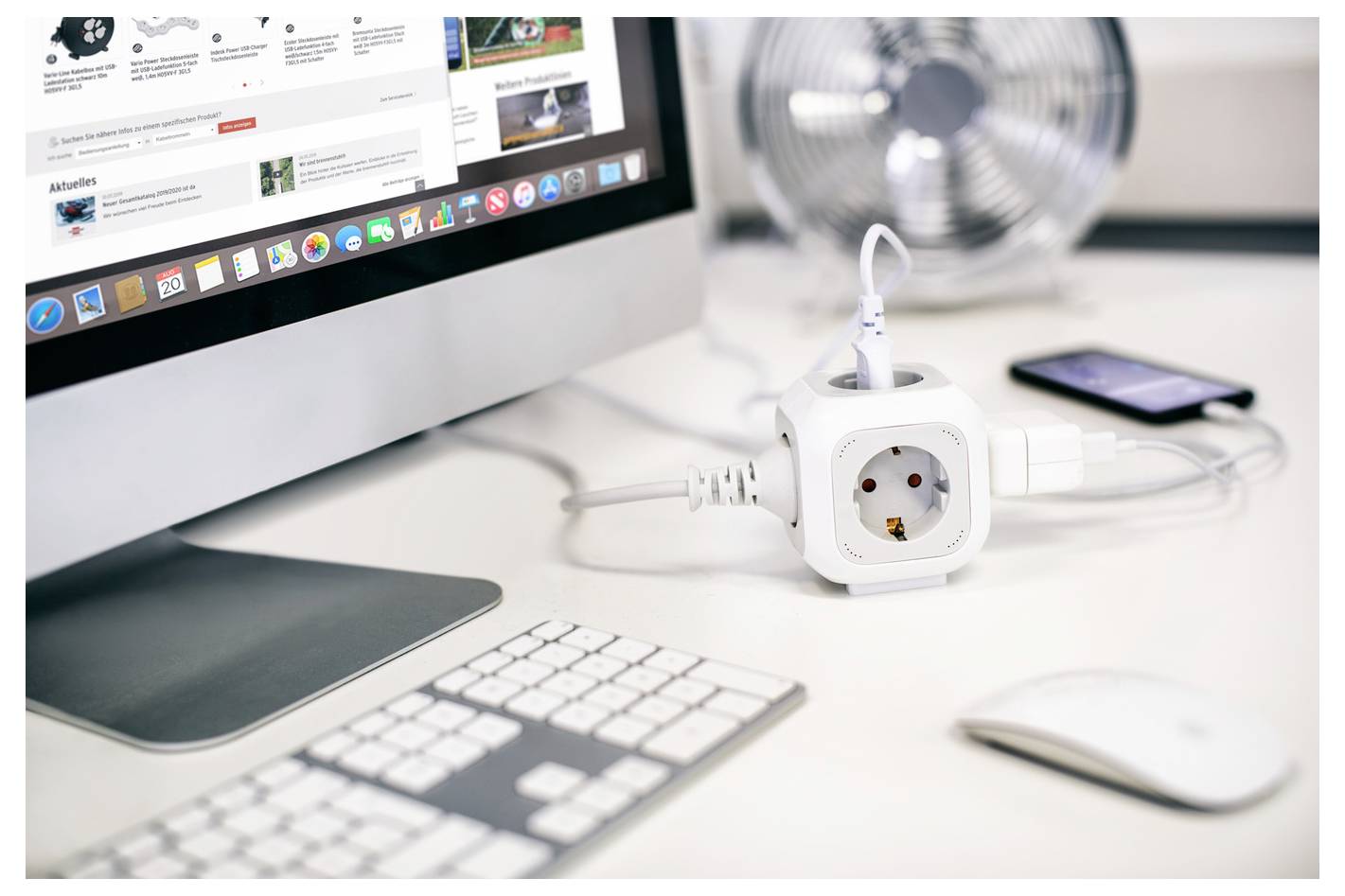 Spazio di lavoro desktop con iMac, tastiera, mouse, adattatore di alimentazione, ventilatore e smartphone su una scrivania bianca