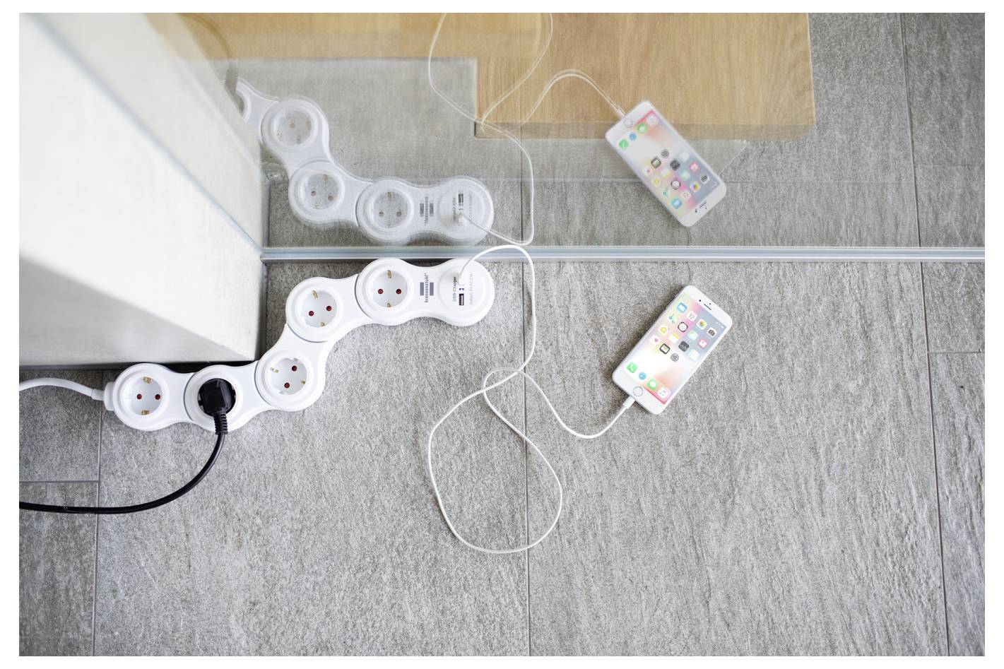 Due smartphone collegati da cavi di ricarica bianchi si trovano su un tappeto accanto a una parete a specchio e a ciabatte elettriche collegate a una presa.