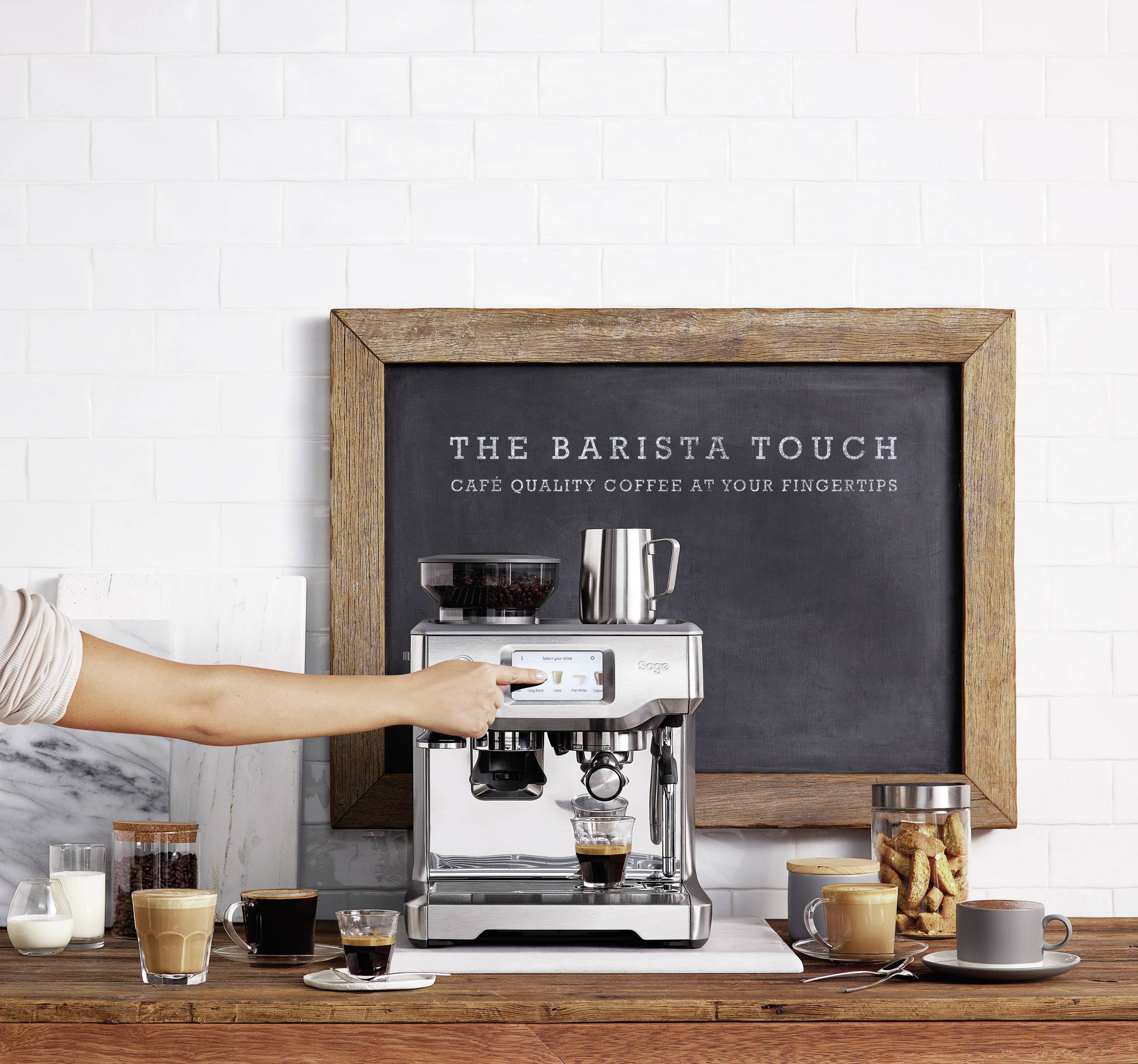 'Macchina per espresso su un piano di lavoro in cucina, circondata da tazze da caffè e bicchieri. Insegna con scritto: The Barista Touch'.