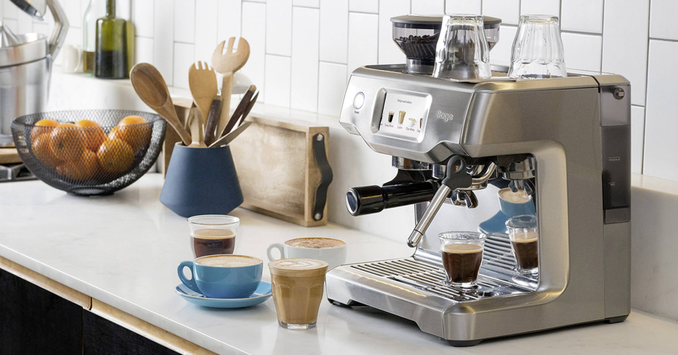 Macchina da caffè espresso su un piano da cucina con caffè preparati nelle tazze. Accanto, un contenitore con utensili da cucina in legno.