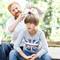 Un adulto pettina i capelli di un bambino con un dispositivo elettrico all'aperto. Entrambi sorridono rilassati durante il processo.