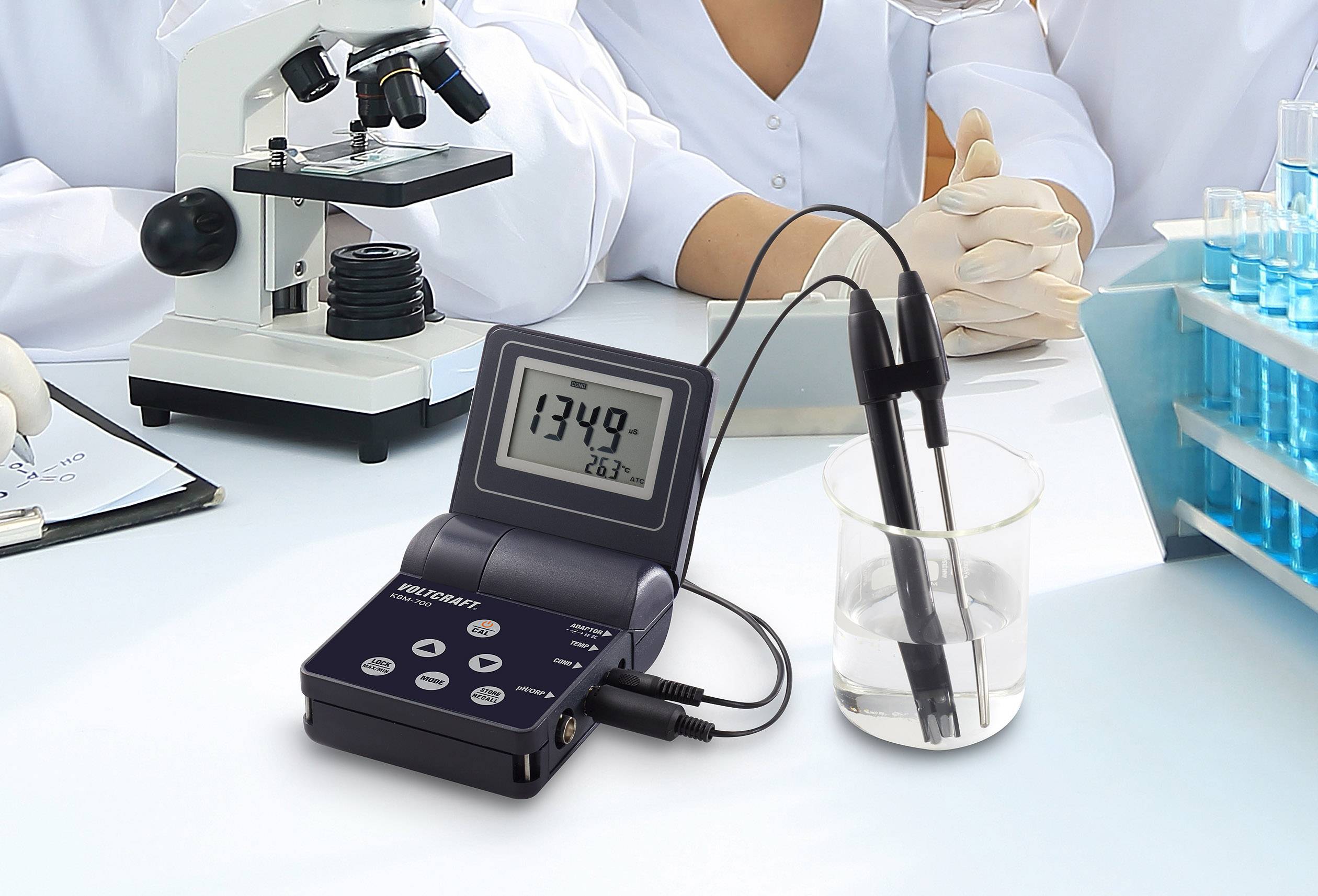 Un misuratore di pH indica il valore 13,49 mentre è immerso in un bicchiere d'acqua. Sullo sfondo un microscopio e scienziati in camice da laboratorio.