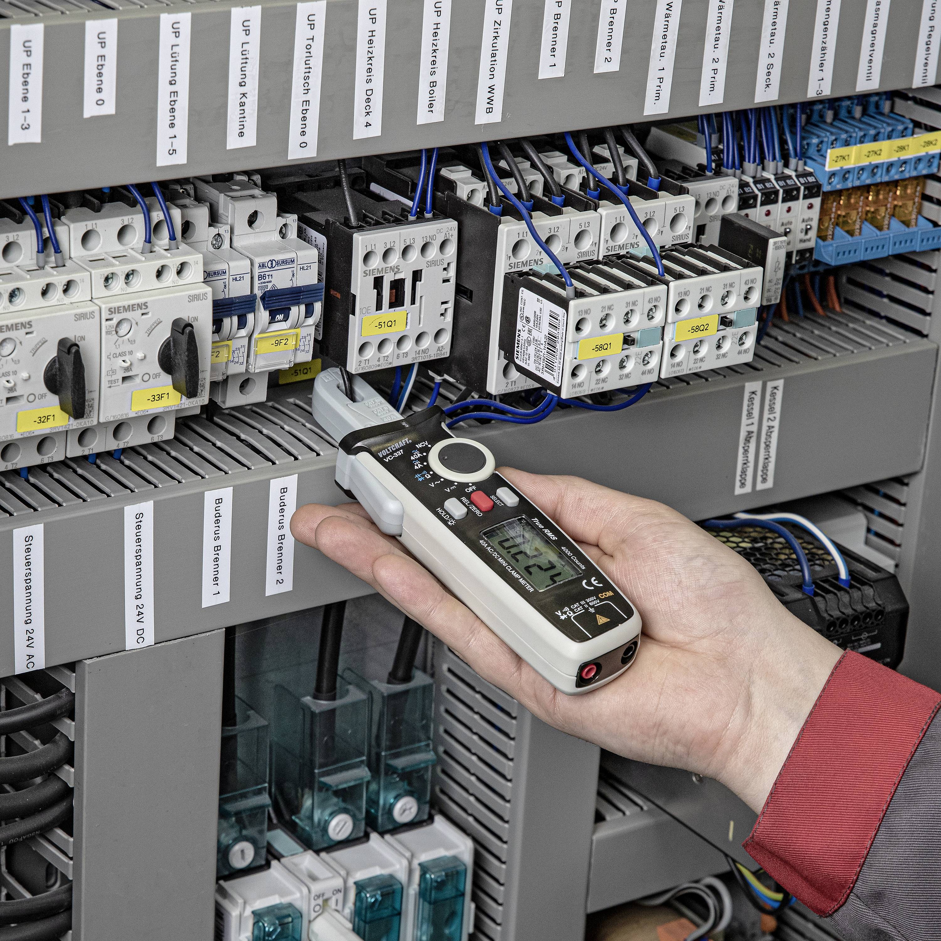 Una mano tiene un multimetro digitale e misura la corrente in un quadro elettrico aperto con diversi cablaggi e dispositivi.