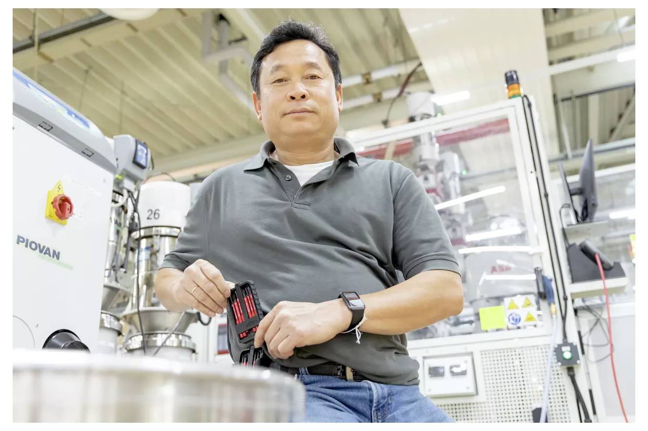 Una persona in un ambiente di fabbrica tiene piccole attrezzature, apparendo concentrata. Macchinari e pannelli di controllo lo circondano, indicando un ambiente industriale.