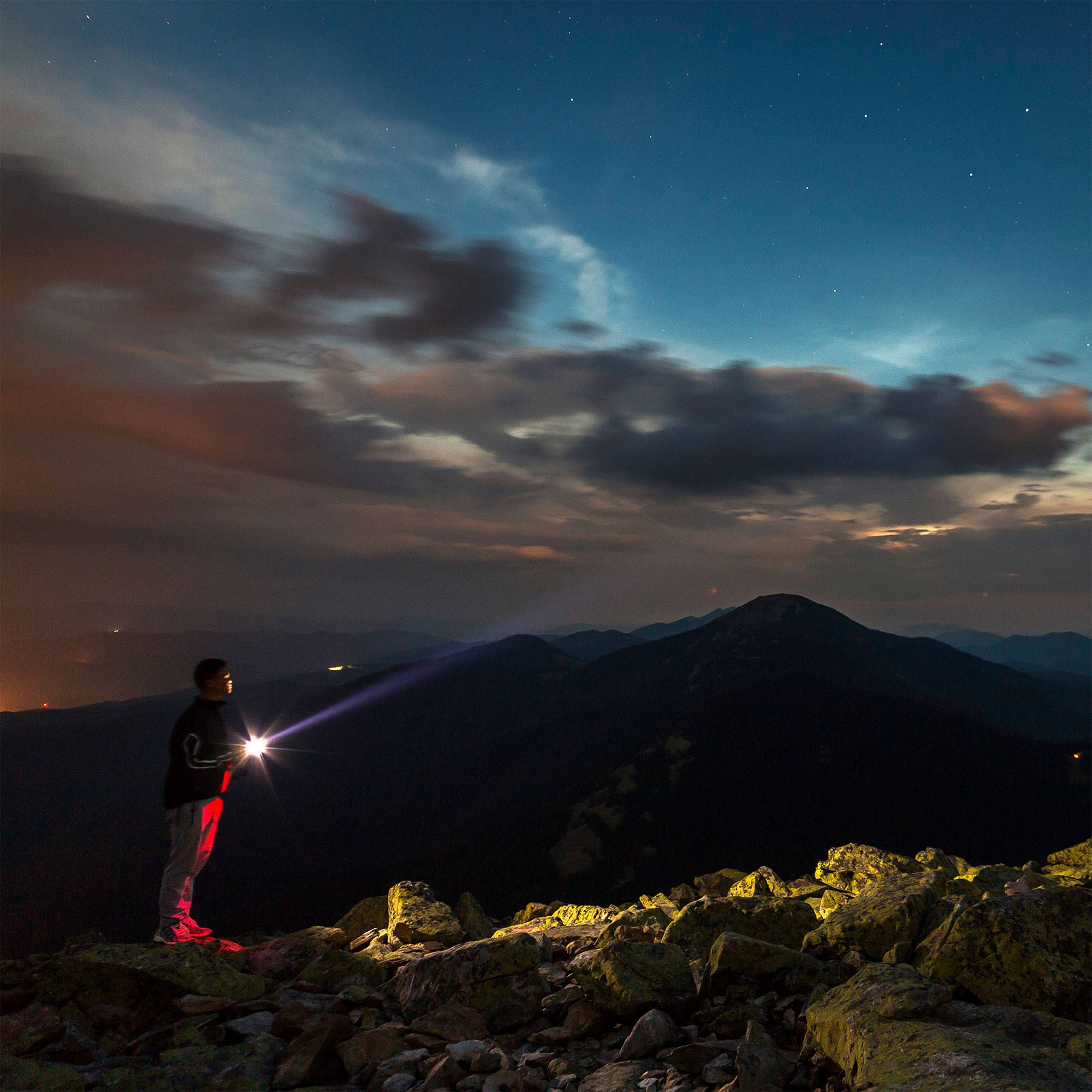 Un uomo è in piedi sulla cima di una montagna di notte e illumina la distanza con una torcia. Il cielo è nuvoloso, con un bagliore azzurro.