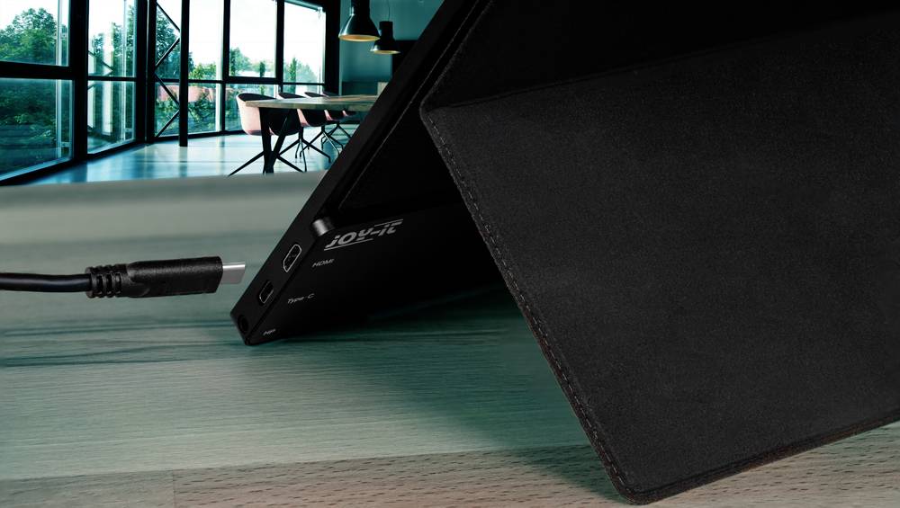 Un notebook viene caricato con un cavo di alimentazione su un tavolo di legno; sullo sfondo si intravedono grandi finestre e mobili per ufficio.