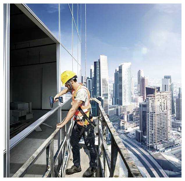 Un operaio edile con indosso l'equipaggiamento di sicurezza installa una finestra su un grattacielo, con la skyline cittadina visibile sullo sfondo.