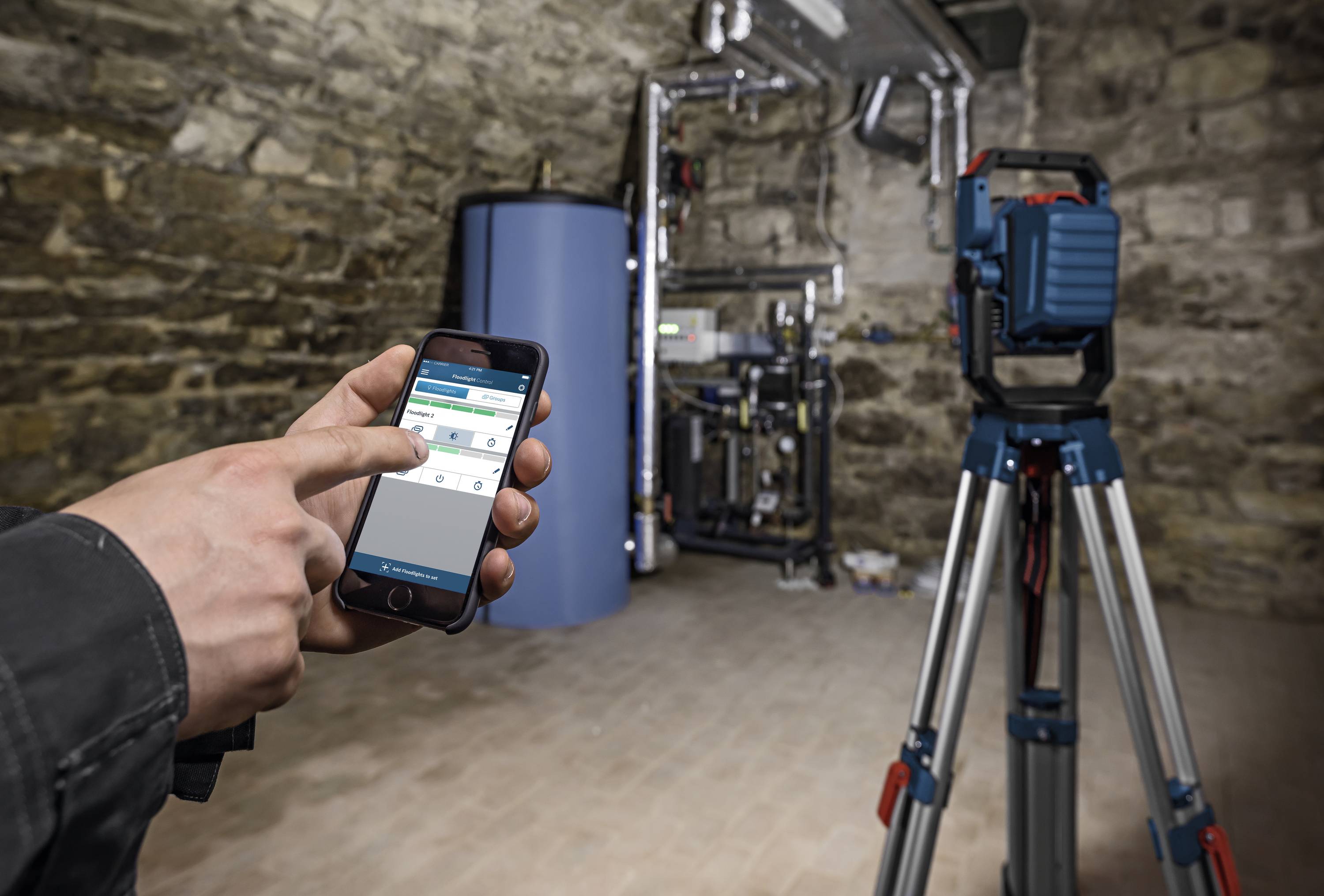 Una persona sta azionando uno smartphone per controllare un dispositivo in un scantinato con pavimento in mattoni. Sullo sfondo è presente un grande dispositivo blu.