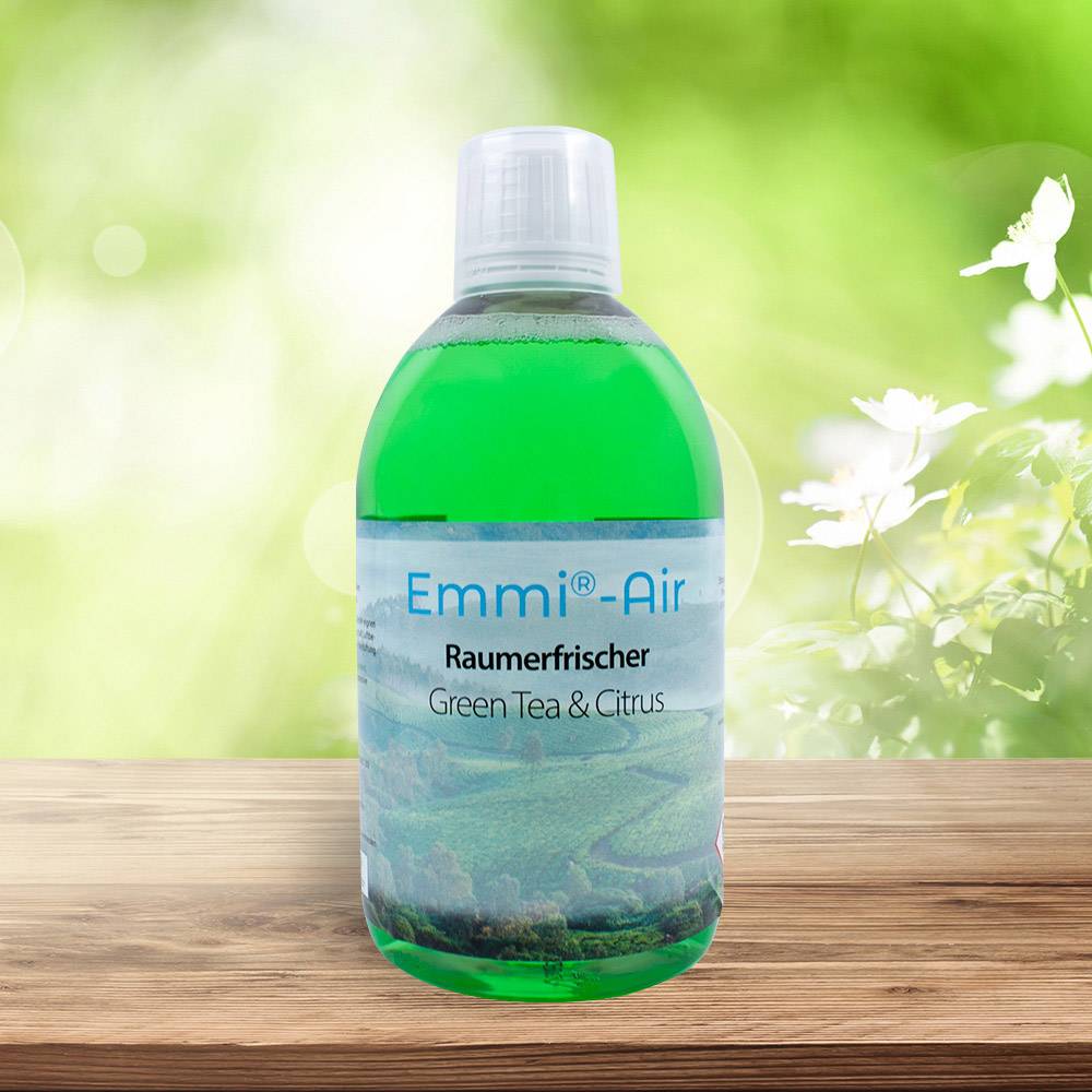 Una bottiglia verde con 'Emmi-Air Deodorante per ambienti Green Tea & Citrus' è poggiata su un supporto in legno, sullo sfondo natura sfocata.