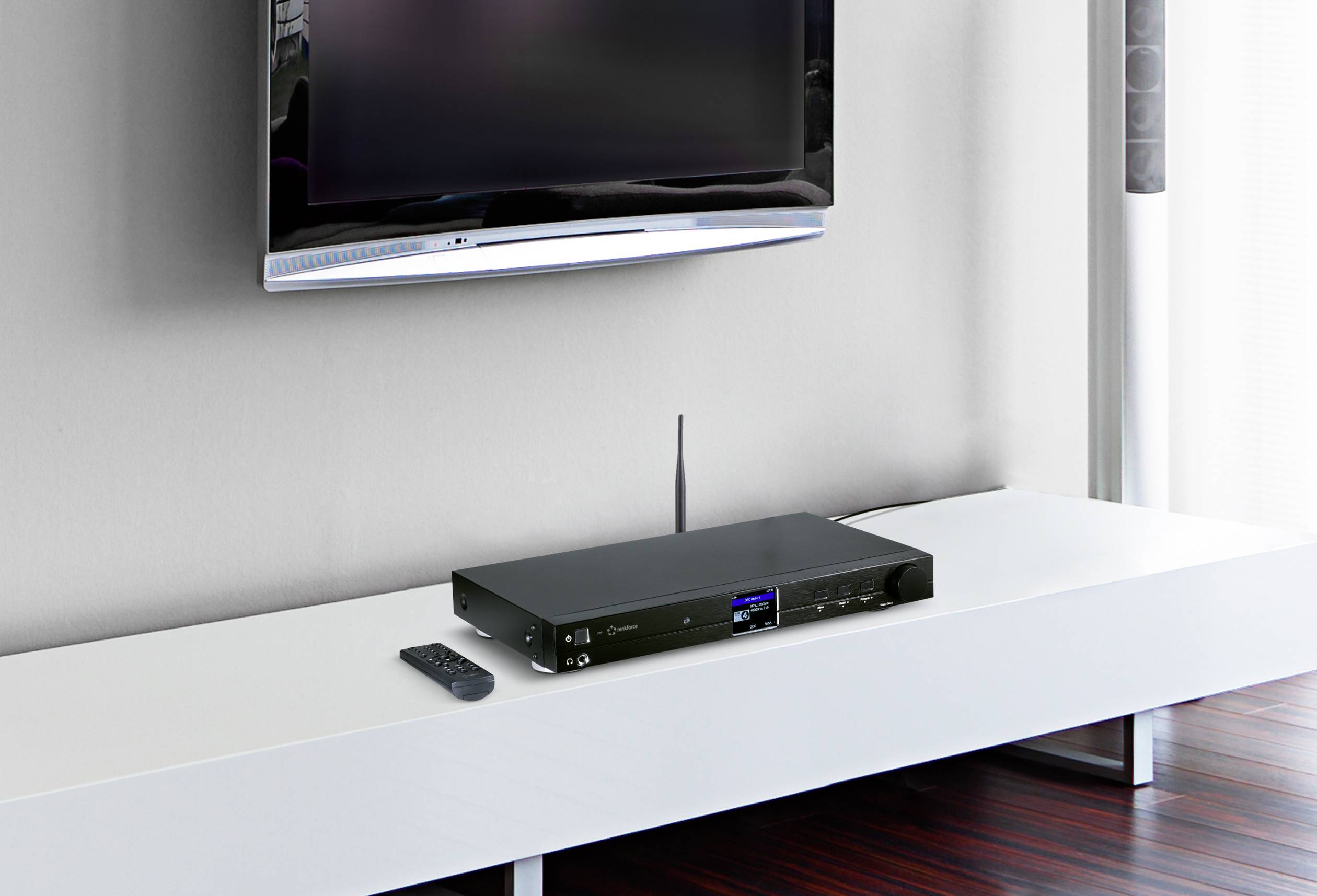 Un ricevitore nero moderno è posizionato su una console TV bianca. Accanto è poggiato un telecomando. Sullo sfondo, un televisore a schermo piatto è montato a parete.