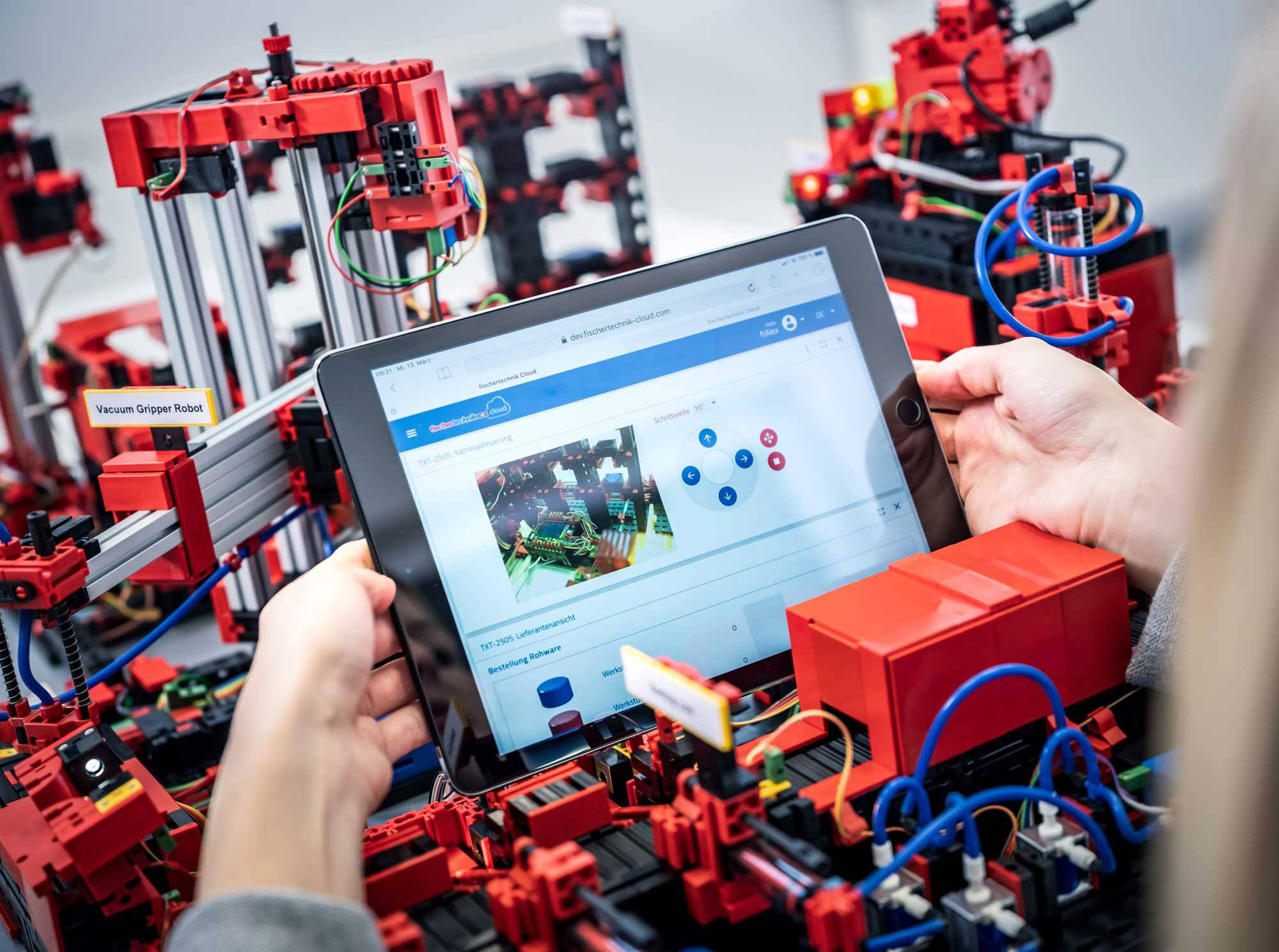 Una persona tiene un tablet tra le mani che mostra il controllo di un complesso sistema robotico rosso con cavi e parti meccaniche.