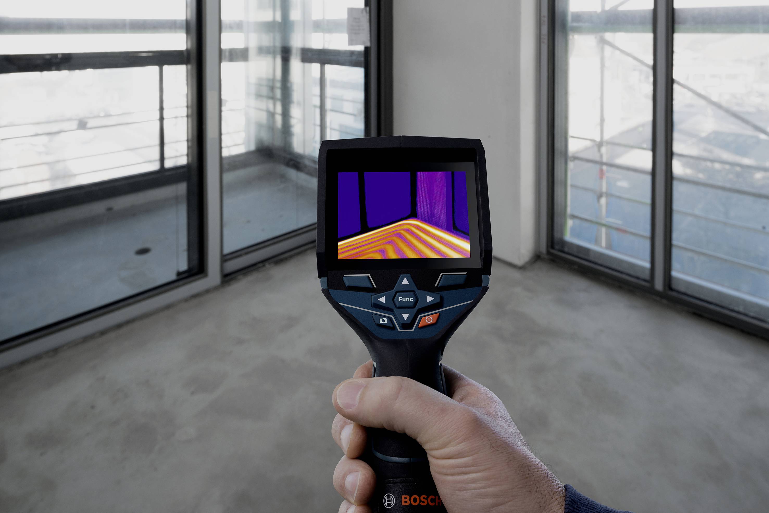 Mano che tiene una termocamera, mostra le differenze di temperatura sul pavimento in una stanza con pareti di vetro. Focus sul display della fotocamera con scala di colori.