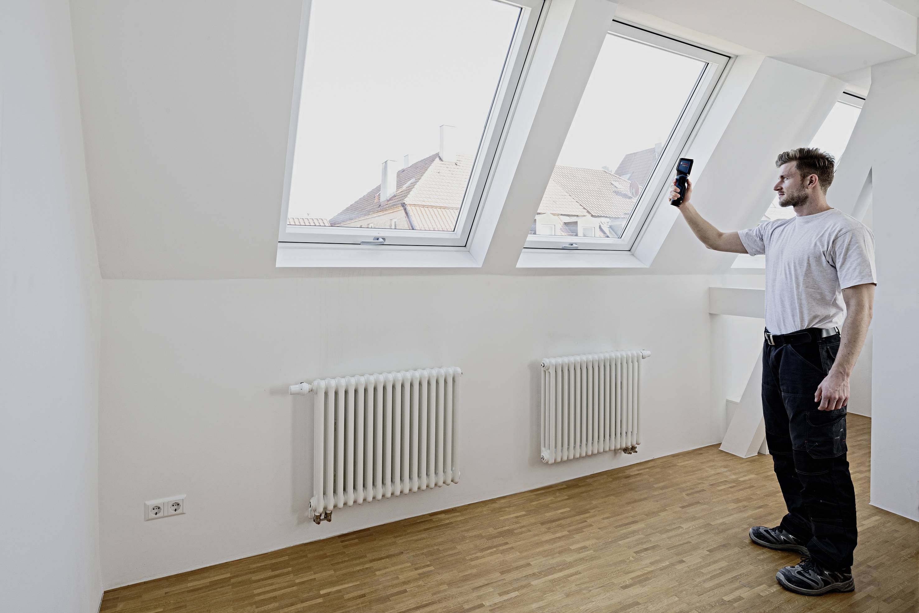 Un uomo in abiti da lavoro è in piedi in una stanza luminosa con due finestre a tetto e due radiatori. Tiene in mano un dispositivo e lo sta osservando.