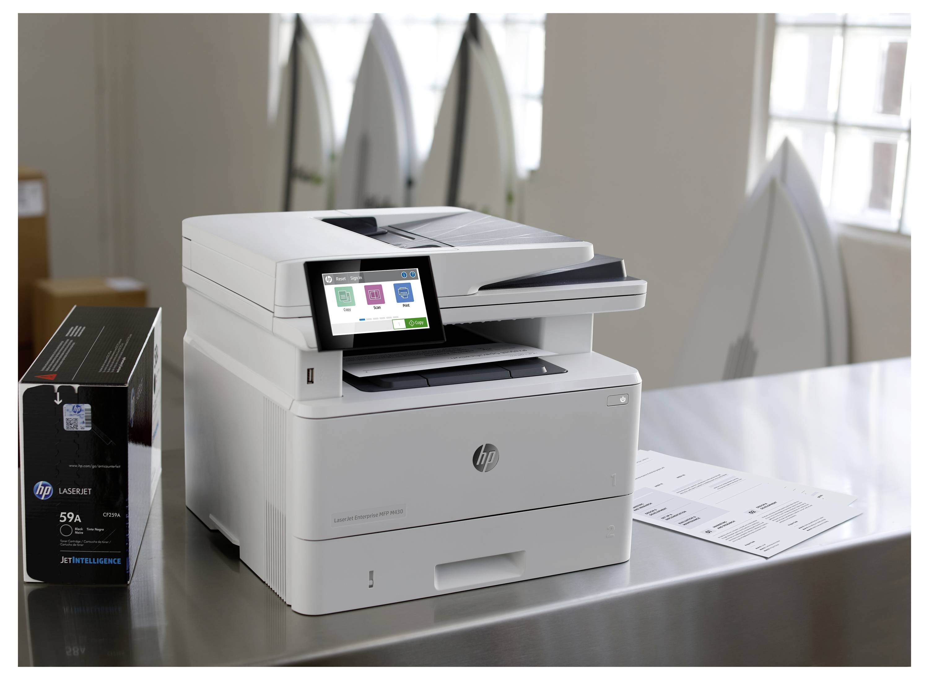 Stampante laser multifunzione HP su un tavolo accanto a una scatola di toner, con display touchscreen e vassoio di uscita della carta in un ambiente d'ufficio.