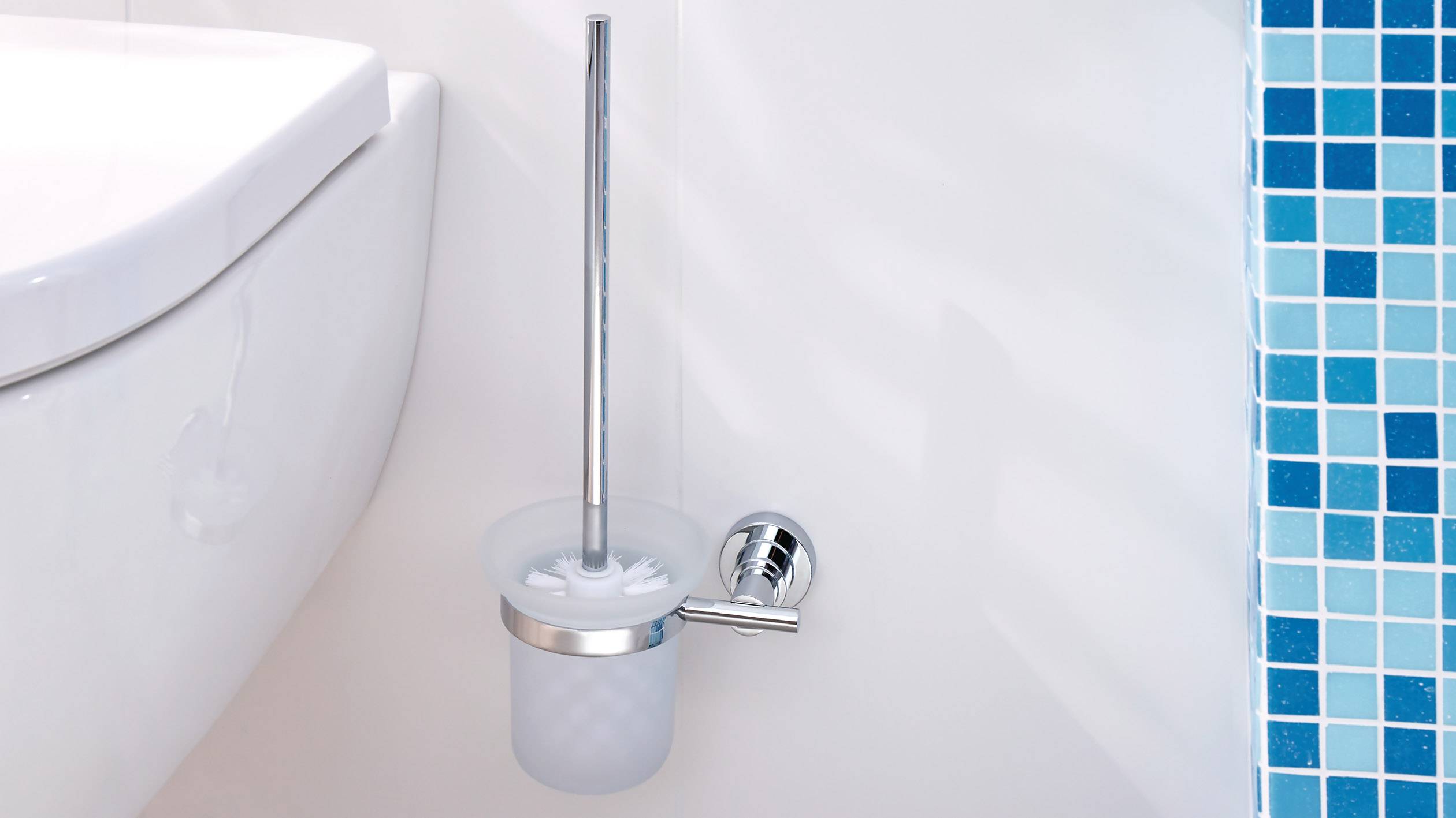 Spazzolino per WC in un supporto accanto a un muro piastrellato, con il bordo di un water visibile a sinistra.