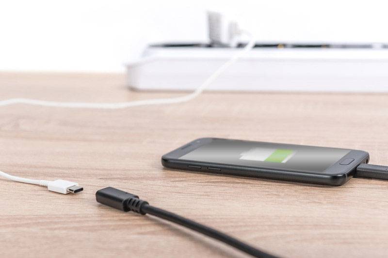 Uno smartphone è poggiato su un tavolo di legno e sta caricando tramite un cavo. Sullo sfondo è visibile una ciabatta elettrica.