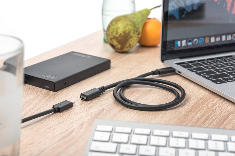 Un laptop su un tavolo con un cavo collegato, accanto a un hard disk esterno. Sullo sfondo ci sono frutta e una bottiglia.