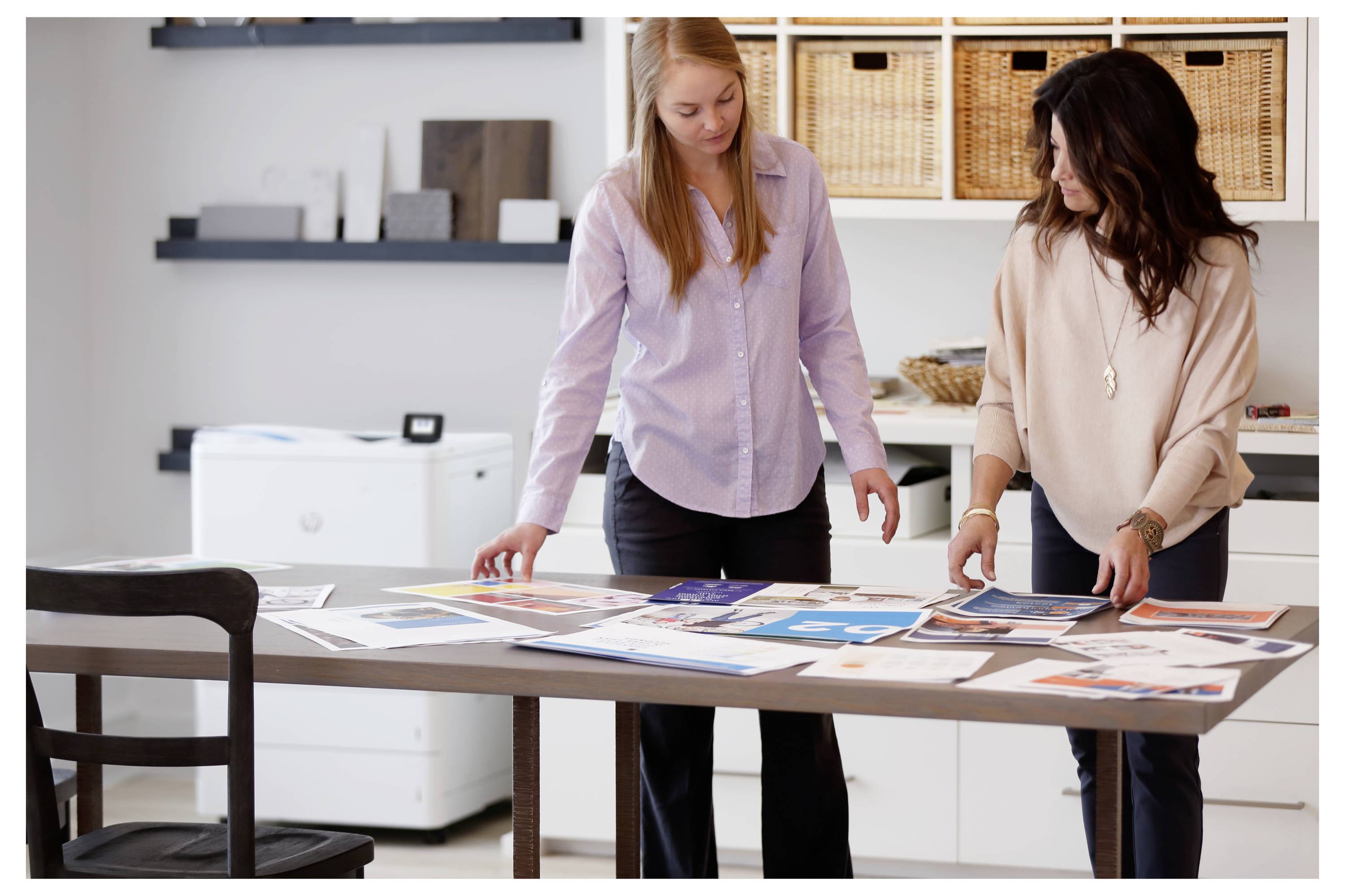 Due donne esaminano layout di design disposti su un tavolo in uno spazio di lavoro d'ufficio luminoso.
