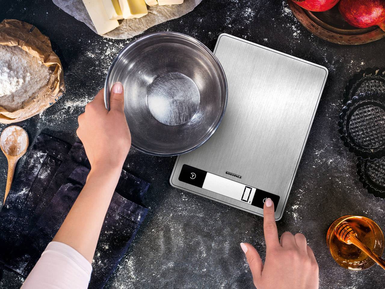 Una persona sta pesando farina in una ciotola su una bilancia digitale da cucina. Circondata da ingredienti da forno su un tavolo.