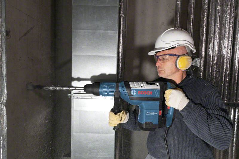 Un operaio con occhiali protettivi, casco e protezioni per l'udito sta utilizzando un grande trapano Bosch per forare un muro in cemento.