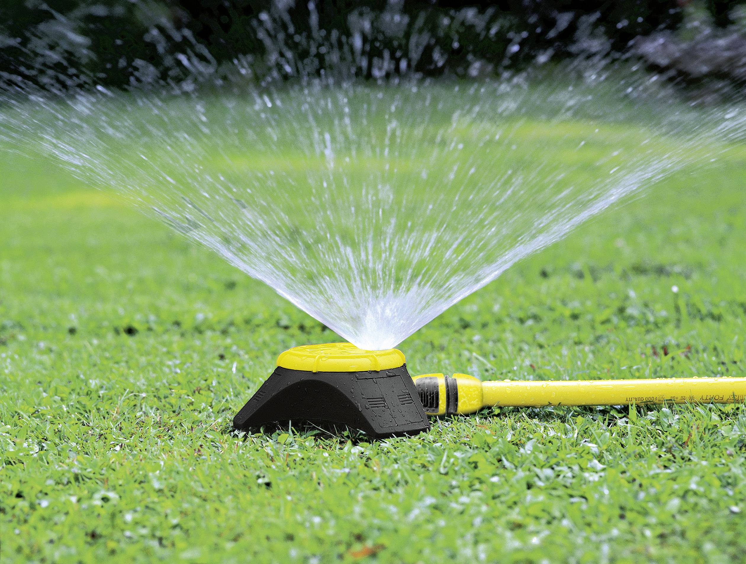 Un irrigatore giallo sta innaffiando l'erba verde nel giardino. Getti d'acqua spruzzano in tutte le direzioni per coprire uniformemente l'area.