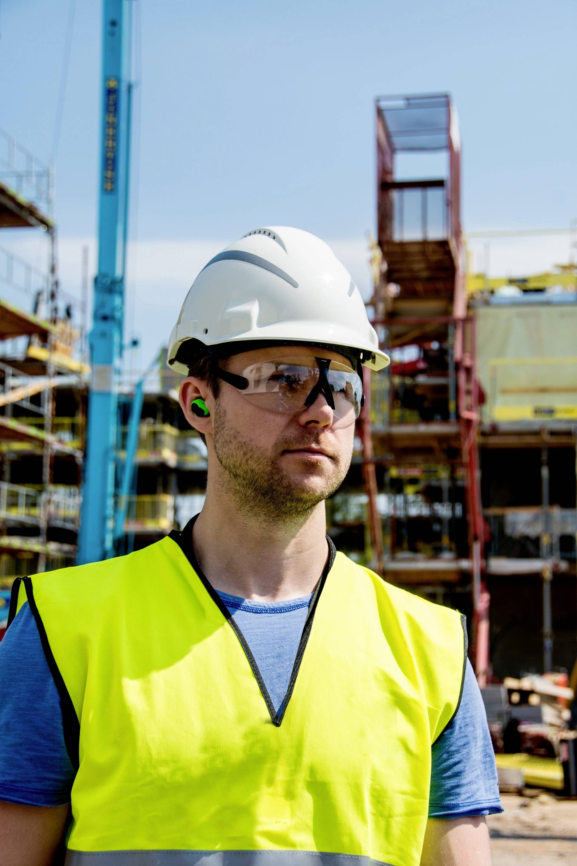 Operaio edile con casco di protezione e giubbotto di sicurezza in piedi davanti a un cantiere in una giornata di sole. Sullo sfondo si vedono ponteggi e gru.