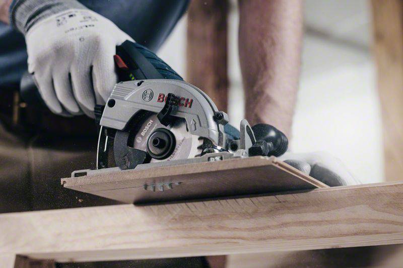 Una persona sta segando una tavola di legno con una sega circolare manuale. La sega è della marca Bosch. La persona indossa guanti da lavoro.