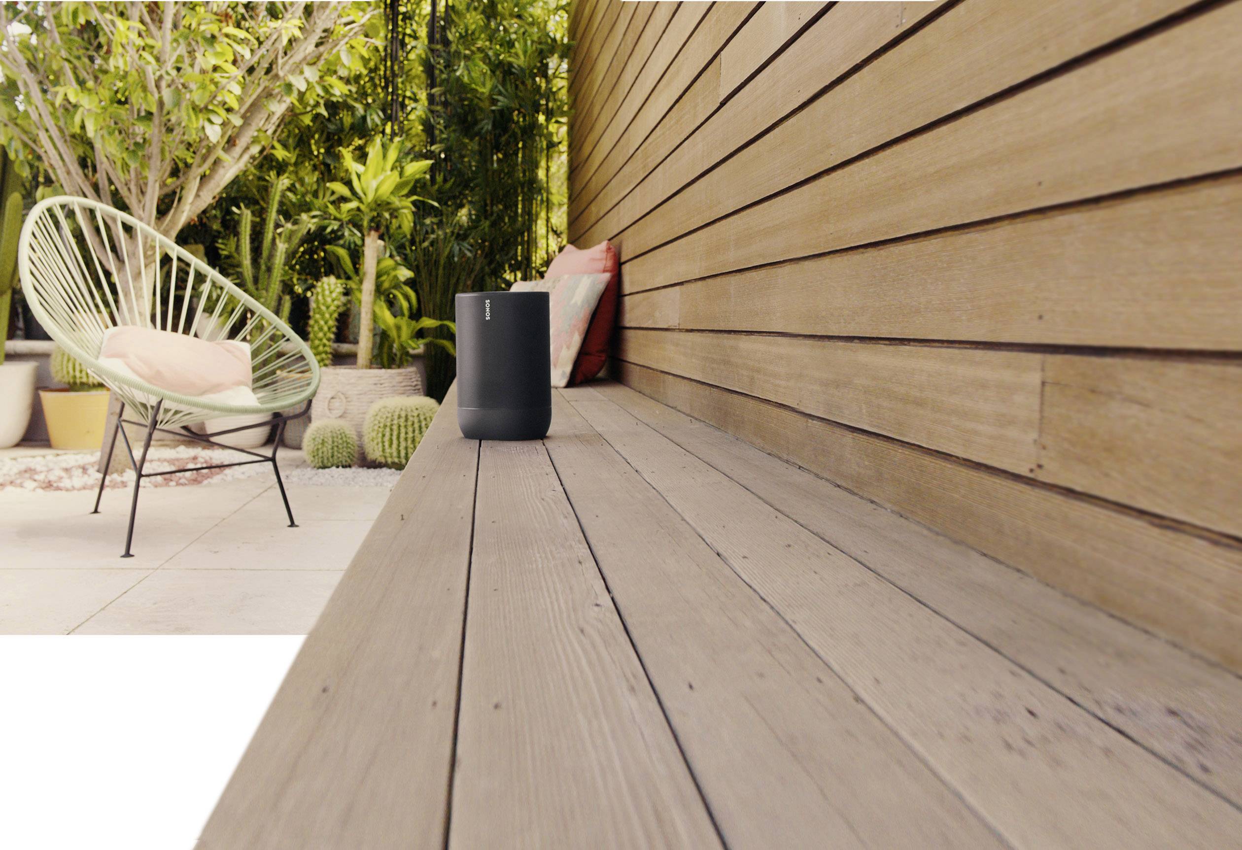 Spazio esterno moderno con rivestimento in legno, una sedia, cuscini e un altoparlante su una lunga panchina di legno. Piante verdi rigogliose sullo sfondo.