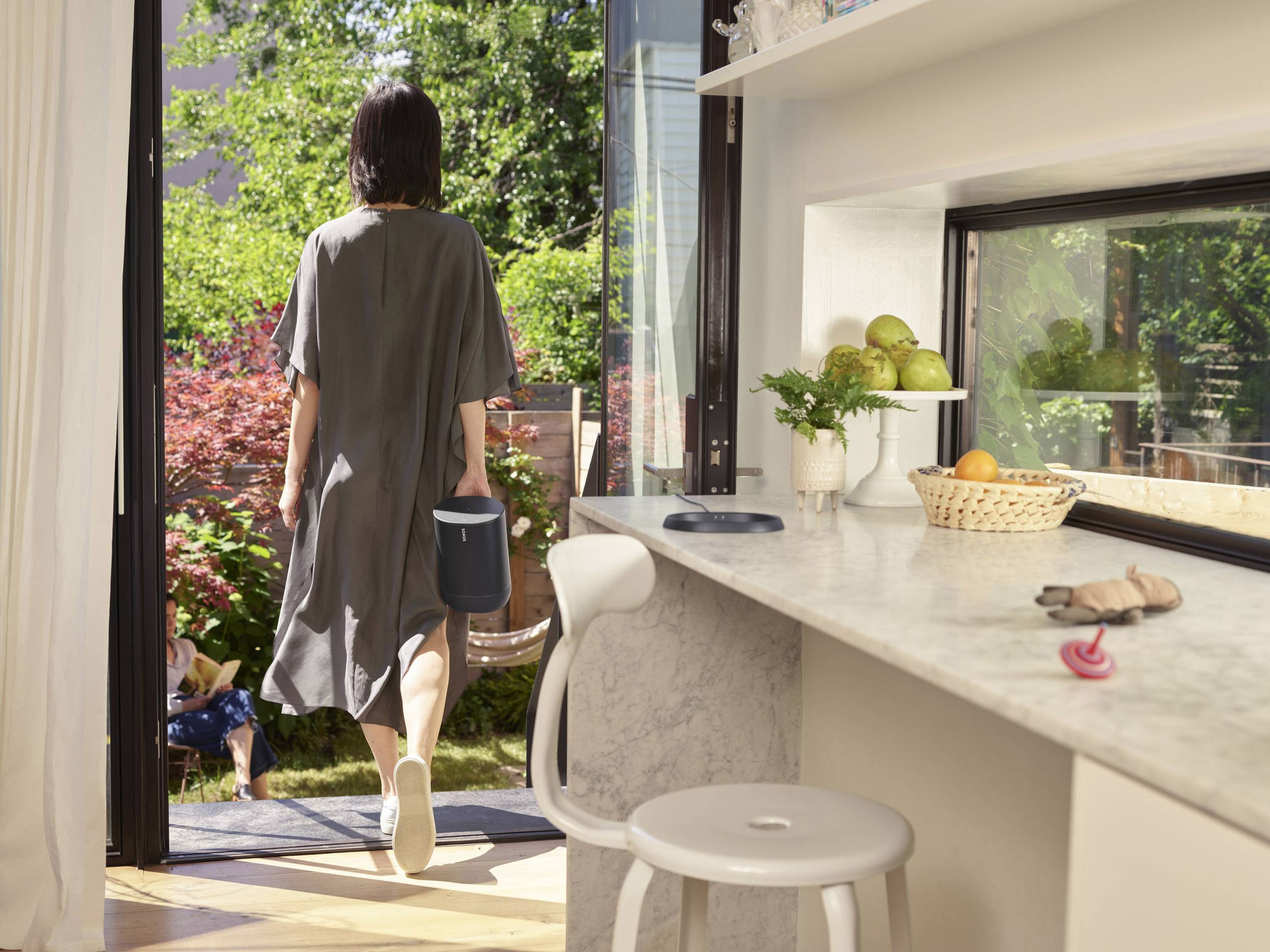 Una donna con un abito grigio esce in un giardino soleggiato. Sul bancone della cucina ci sono un cestino di frutta e un telefono.