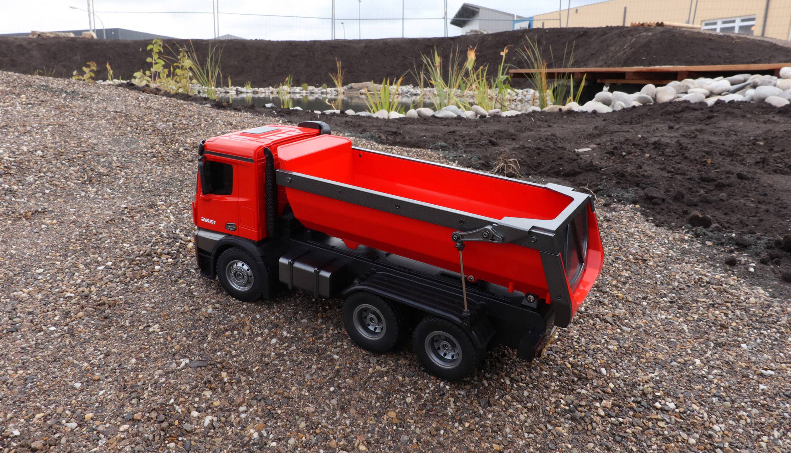 Un camion giocattolo rosso è fermo su un terreno di ghiaia davanti a un terrapieno. Sullo sfondo si vedono piante e un edificio.