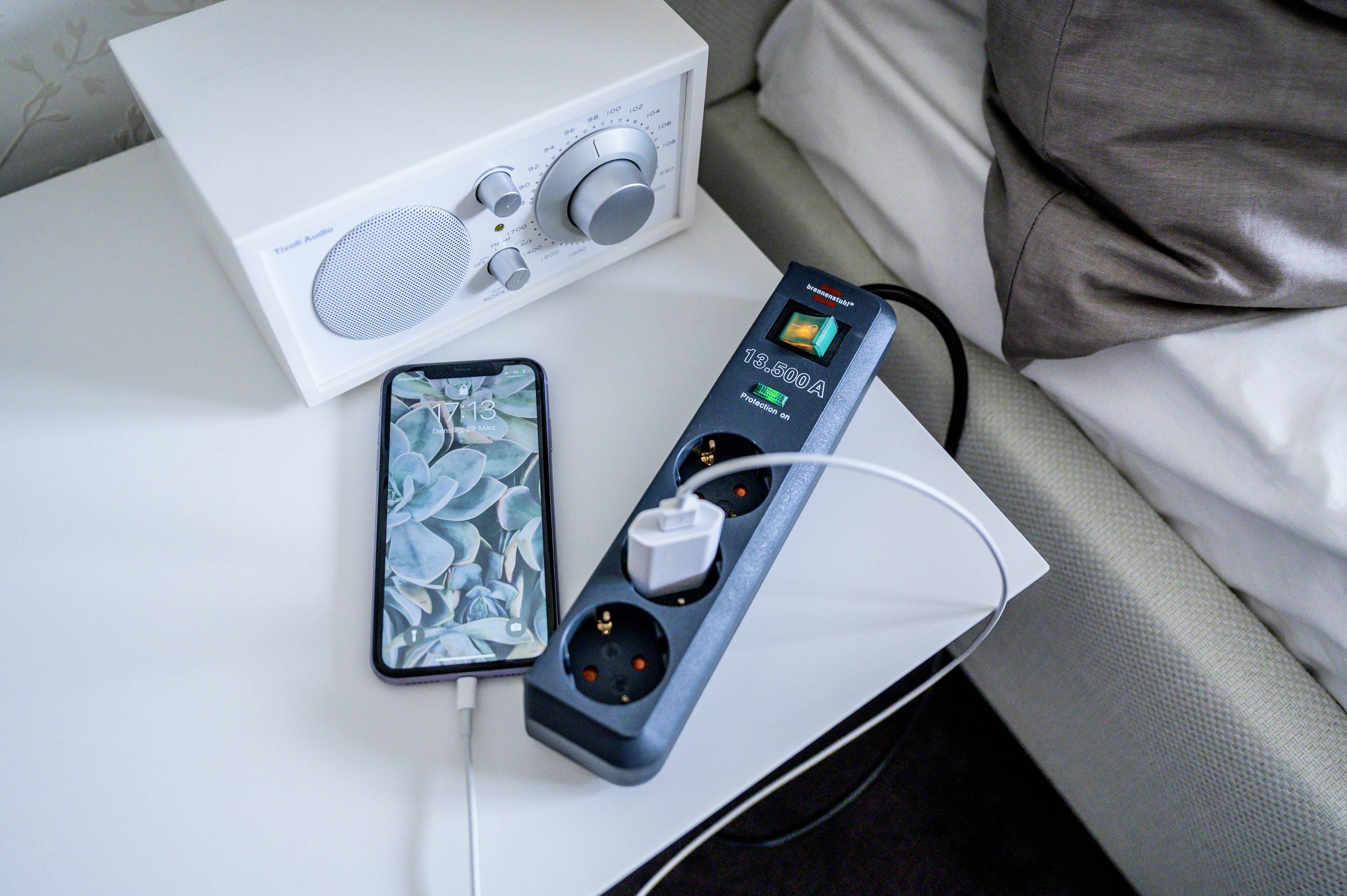 Un smartphone collegato tramite un cavo di ricarica a una presa multipla, accanto a un radio su un comodino.