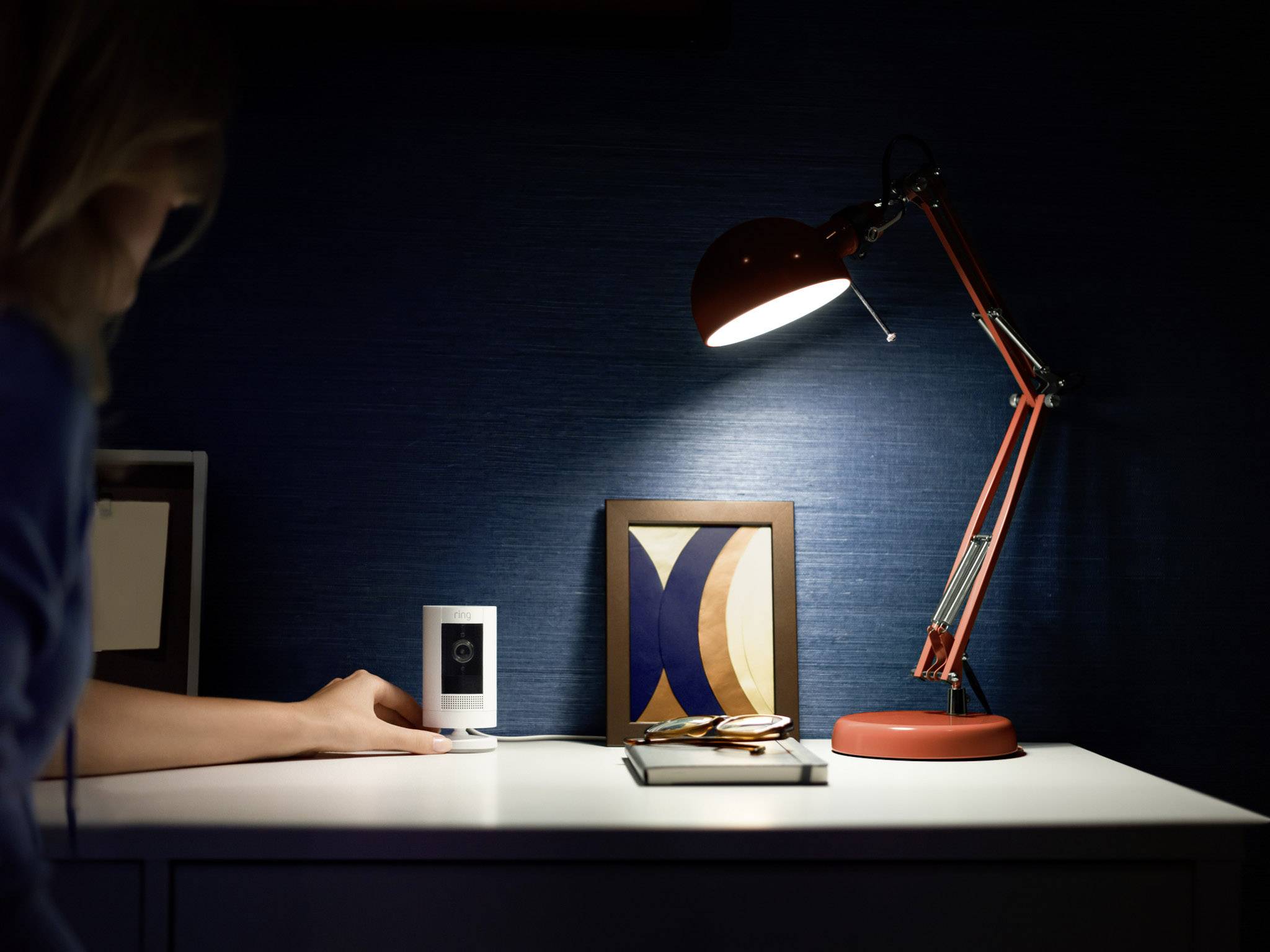 Una persona sta per afferrare un dispositivo su un tavolo illuminato da una lampada da tavolo rosso-arancione. Sullo sfondo si vedono cornici di foto.