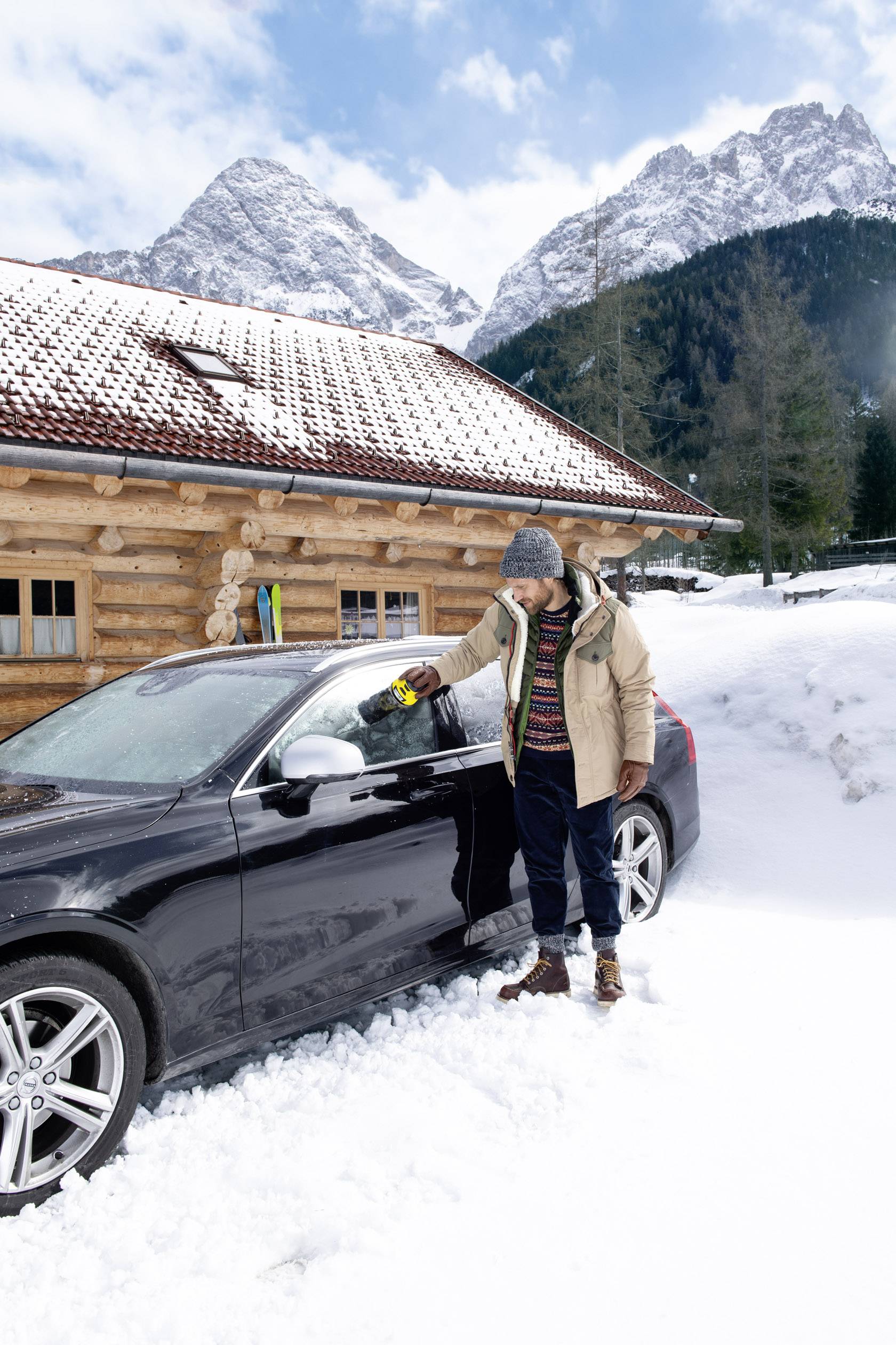 Una persona vestita con abiti invernali rimuove la neve da un'auto nera davanti a un chalet di legno in un paesaggio montano innevato.