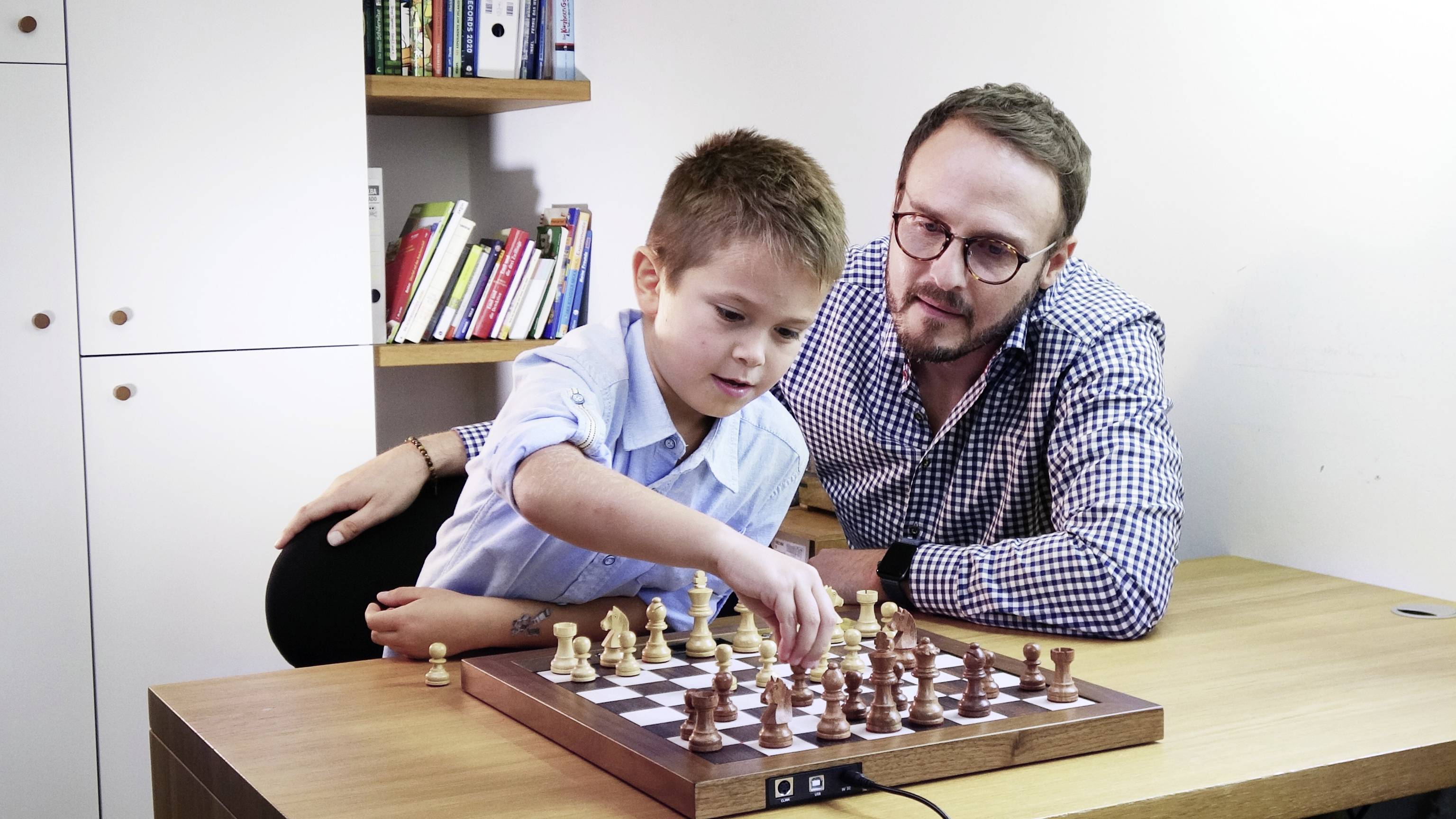 Un bambino gioca a scacchi con un adulto a un tavolo. Si concentrano sul gioco. Sullo sfondo ci sono scaffali di libri.