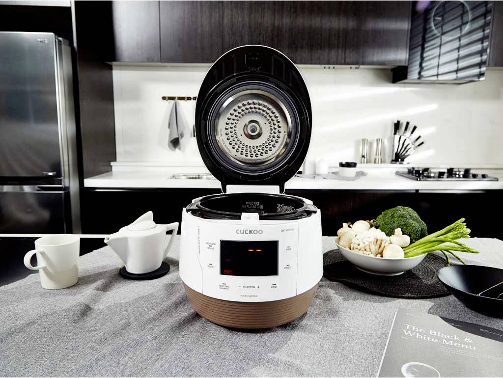 Macchina per il riso su un tavolo da cucina con il coperchio aperto, circondato da verdure, un piatto e una tazza. Cucina moderna sullo sfondo.