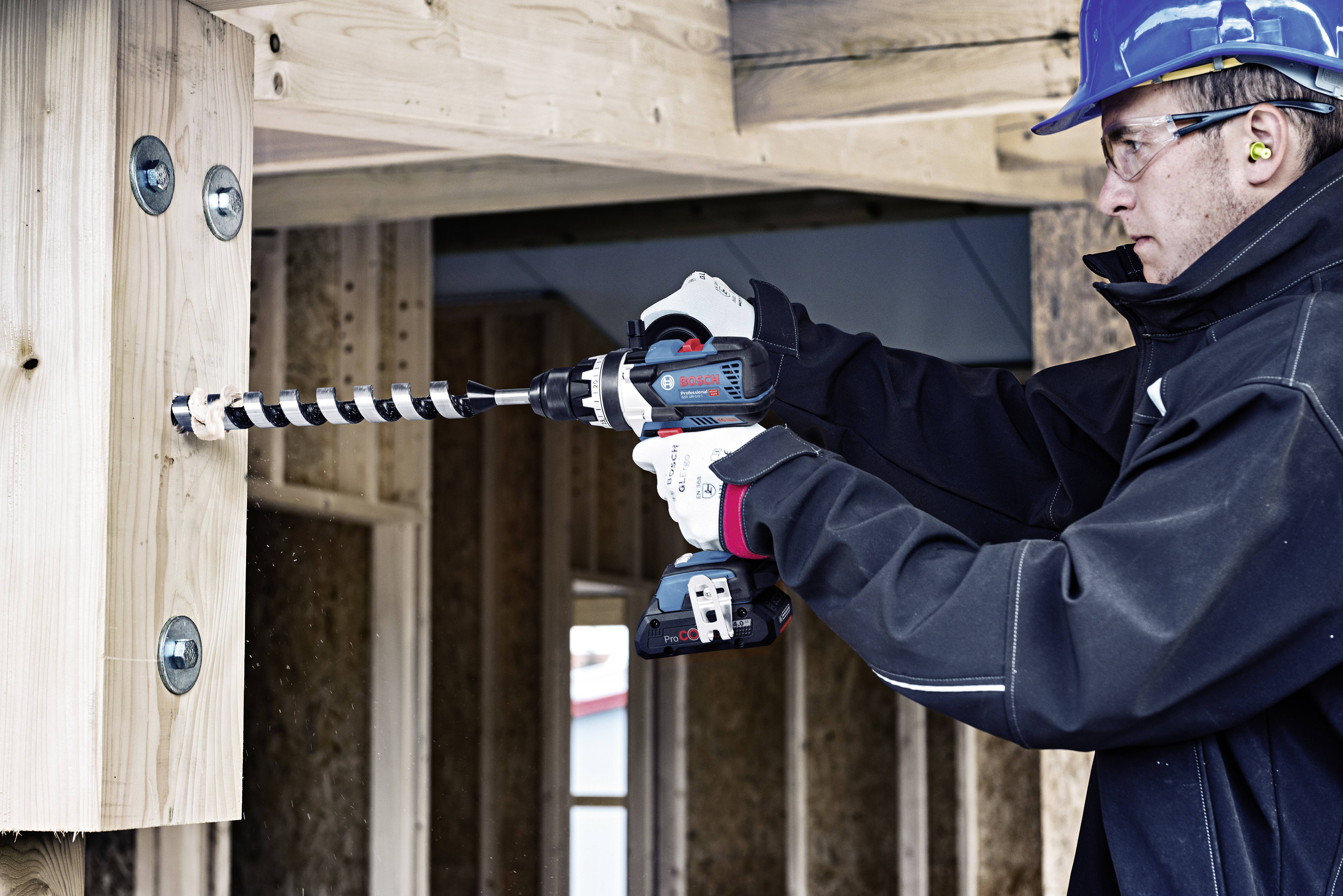 Un lavoratore con casco e dispositivi di protezione sta utilizzando un trapano elettrico per forare un grande foro in una trave di legno.