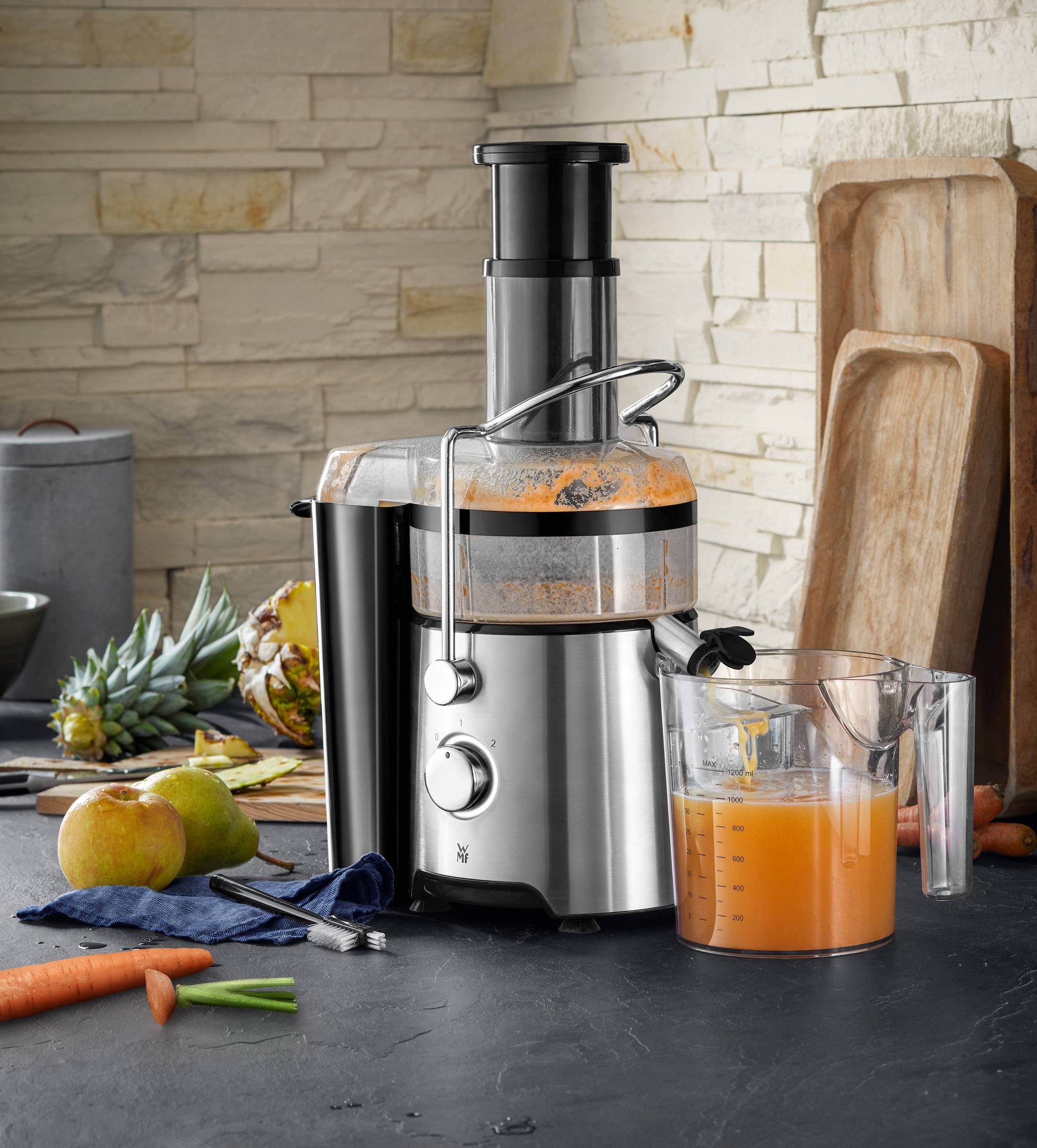 Estrattore su un piano da cucina con succo fresco in una caraffa di misurazione, circondato da frutta e verdura come ananas, mele e carote.