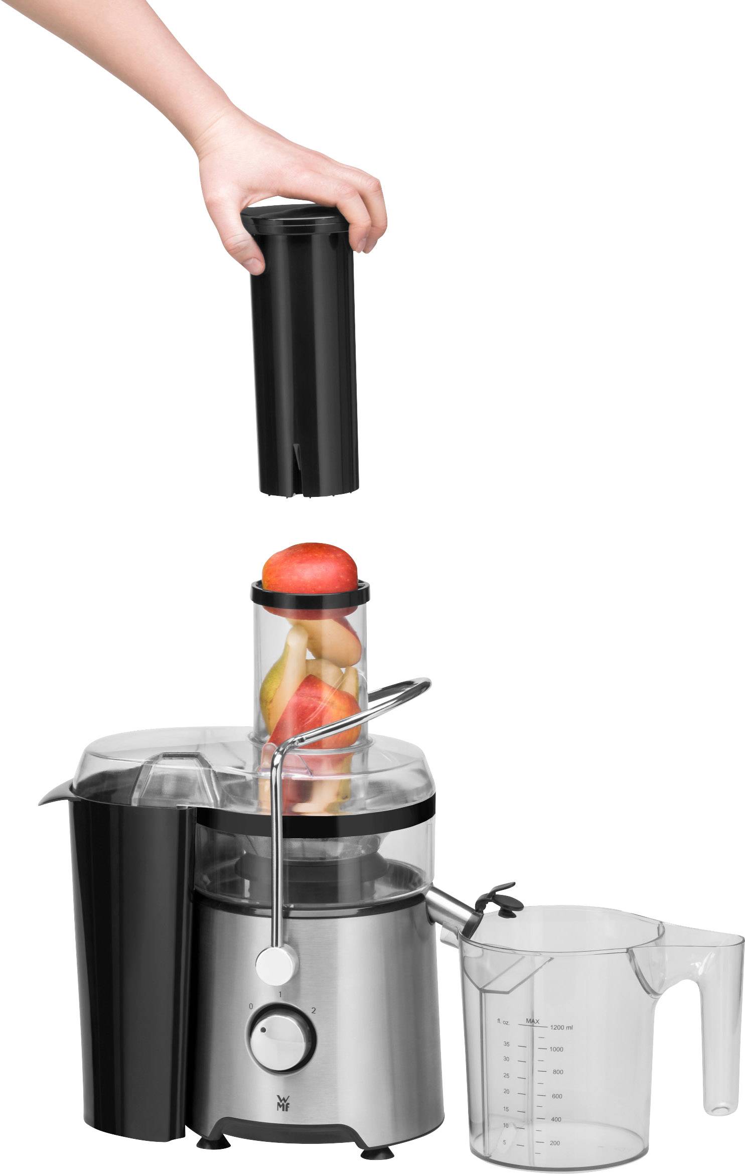 La mano posiziona il coperchio sull'estrattore, riempito con pezzi di mela e pomodoro. Accanto è presente un misurino per raccogliere il succo.